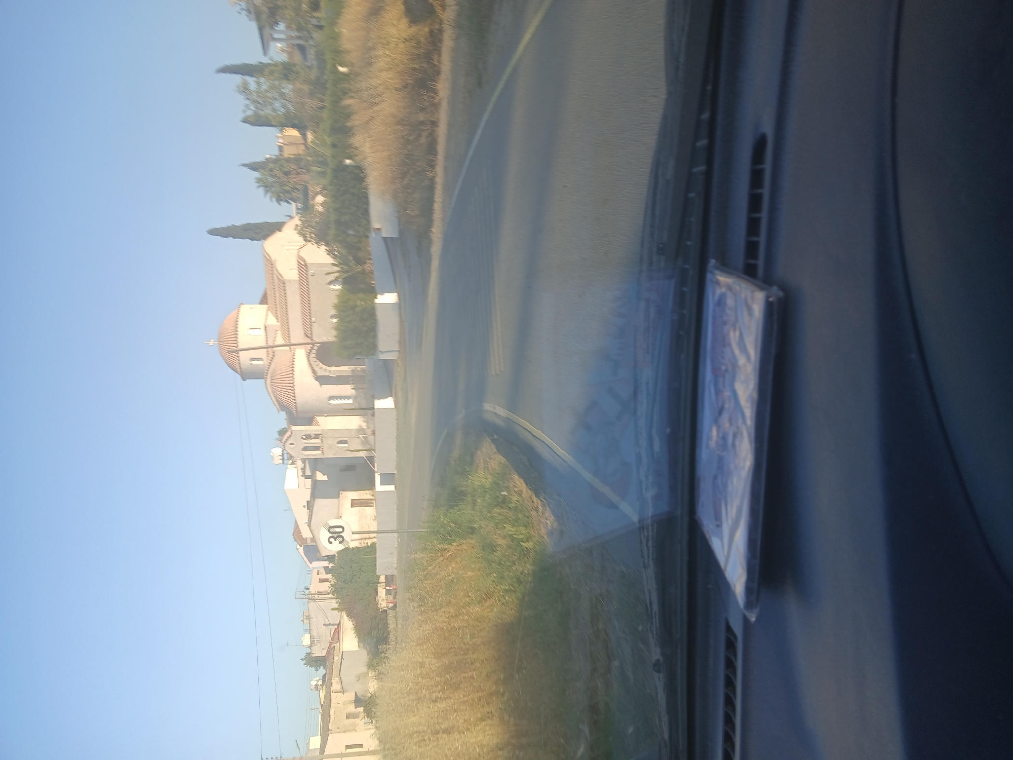 The image presents a sunlit roadside scene in Pera Chorio, Cyprus, viewed from inside a vehicle. The perspective is captured through the front windshield, with a dark dashboard and a reflective object, possibly a CD case, visible on the right side of the frame.
Outside the vehicle, a paved road with a yellow dividing line curves gently to the right. The left side of the road is bordered by stretches of dry, golden-brown grass and some green vegetation, including tall, slender trees. Under a clear, bright blue sky, several buildings are visible.
The most prominent structure is a light-colored building featuring a distinctive reddish-ttiled dome surmounted by a cross, characteristic of an Orthodox church. Other residential or commercial-style buildings are also present along the road. A white circular speed limit sign with the number "30" in black is clearly visible on a pole beside one of the buildings.
The scene depicts clear, sunny daytime conditions. No people are visible. The overall setting suggests a quiet, possibly rural or semi-urban area in the Mediterranean region.