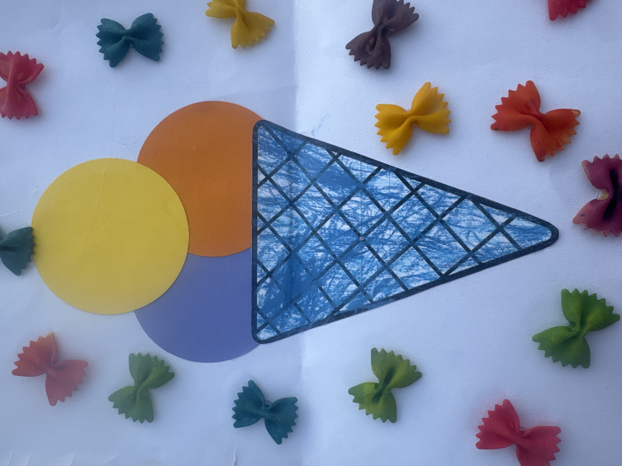 This is a top-down, eye-level, medium shot of an art project on a white background. The art project depicts an ice cream cone with three scoops and a waffle cone. The scoops are colored yellow, purple, and orange. The waffle cone is drawn in blue crayon with a black outline. Surrounding the ice cream cone are numerous colorful bowtie pasta pieces. These pasta pieces are red, green, yellow, blue, brown, and purple. The background appears to be white paper, possibly on a table or wall. The lighting is bright and even. There are no people or animals present in the image. The image does not contain any information that would allow for its location to be identified in Saint-Claude, France.