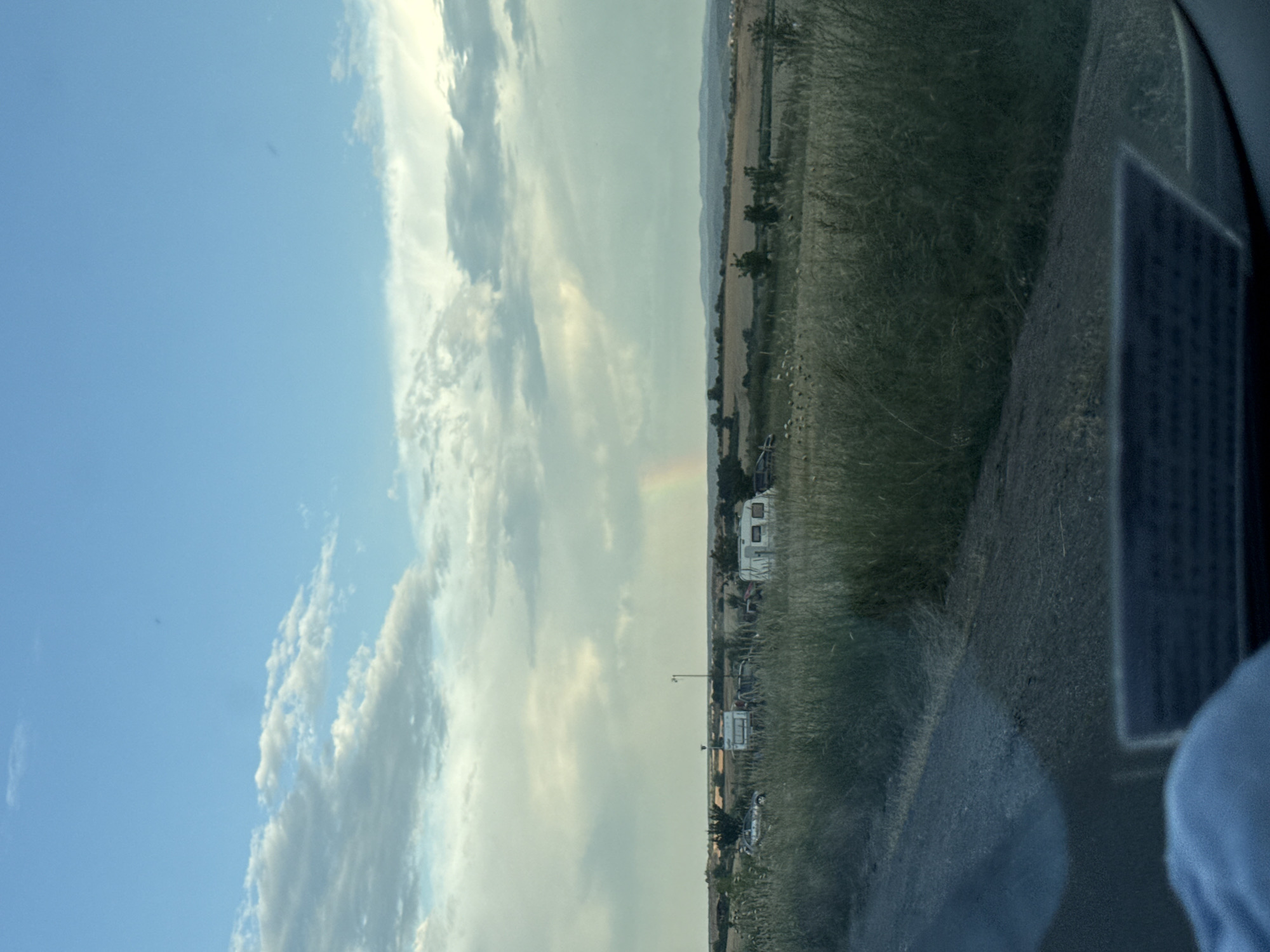 A wide-angle, outdoor, eye-level shot captures a rural landscape under a partly cloudy sky with a faint rainbow arc. The foreground is dominated by a dusty, grey road on the left, with tall, dry grass bordering it on the right. Beyond the grass, a collection of caravans and vehicles are parked in a field. The middle ground shows more fields and scattered trees, leading to a distant line of rolling hills on the horizon. The sky is a mix of bright blue and diffused grey clouds, with the sun's rays creating a subtle glow near the rainbow. The overall impression is of a serene, open space, possibly a campground or rural gathering, located near Tvarditsa, Bulgaria.