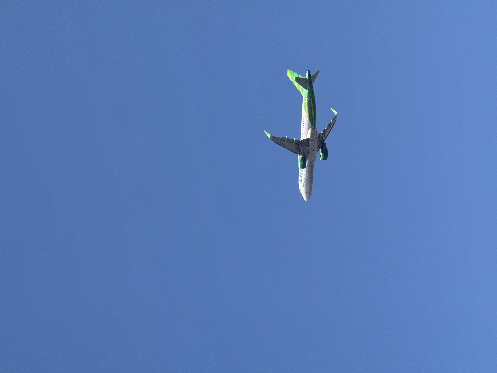 An airplane with a green and yellow livery is captured in flight against a clear blue sky. The aircraft appears to be a commercial jetliner, with its engines mounted under the wings and its tail fin featuring a distinctive green stripe pattern. The nose of the plane is white, and the fuselage is predominantly white with green accents and lettering. The wings are extended, and the landing gear is retracted, indicating it is airborne. The image is a vertical shot, emphasizing the vastness of the sky above. While the specific location of South Denpasar, Indonesia, is mentioned, there are no visual cues in the image itself to confirm this geographical context, such as landmarks or ground features. The scene is solely focused on the airplane in flight.