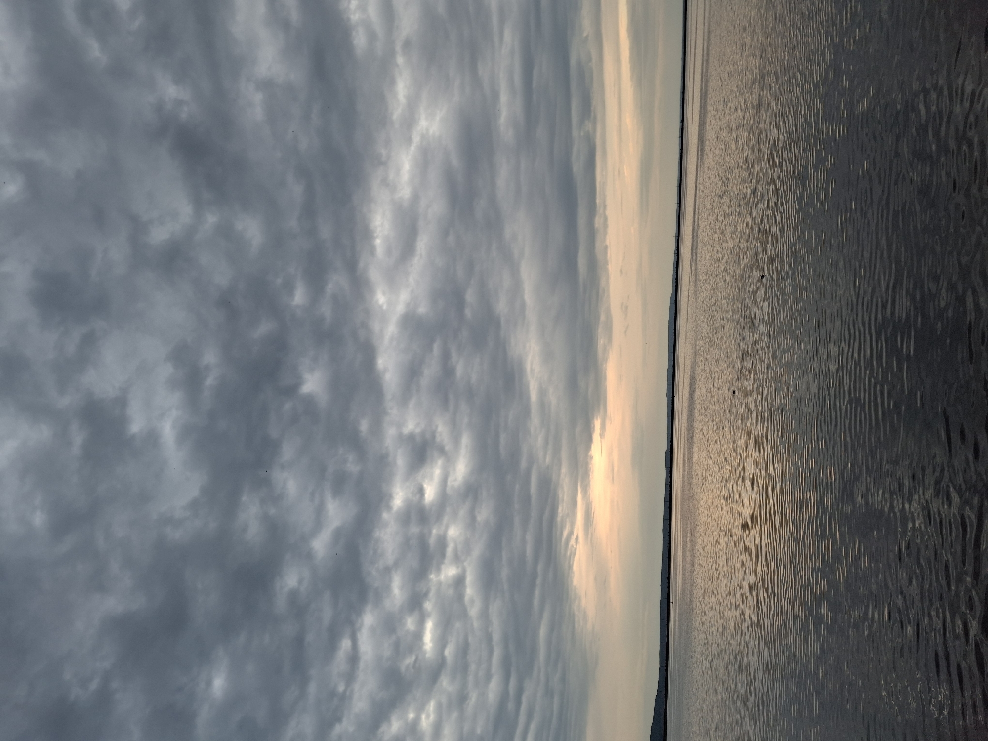 The image shows a horizontal view of a body of water reflecting sunlight, juxtaposed against a cloudy sky. The sky occupies the left side of the frame, filled with layers of gray and white clouds. The water on the right side of the frame shows small ripples and a bright reflection. The location is likely Chaikovsky, Russia, given the image was taken there, as it is situated on the Kama River which could be the body of water visible. There are no people visible in the image, no objects are recognizable besides water and clouds. The only action is the reflection of sunlight on water.