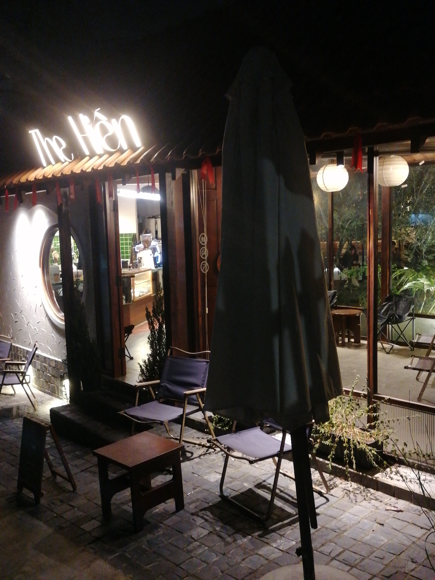 This image depicts an outdoor seating area and entrance to a establishment identified by a neon sign reading "The Hien". The scene is lit by artificial lights, creating a warm and inviting ambiance. The establishment appears to be a cafe or restaurant, with a barista station visible through the glass entrance. A large umbrella dominates the foreground, partially obscuring the view of the seating area, which includes several chairs and small tables. The ground is paved with interlocking bricks, and several potted plants add a touch of greenery to the scene. The overall impression is one of a cozy, nocturnal gathering space.
