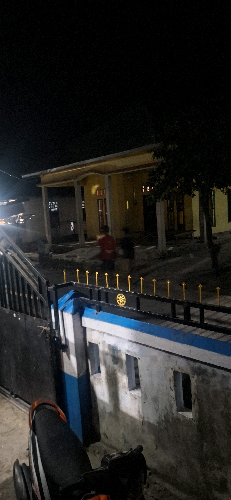 In Desa Molona, Indonesia, at night, a low-angle shot captures a yellow-painted building with visible pillars and red-framed windows, illuminated by interior lights. A person in a red shirt stands in the foreground, near another blurred figure. A fence with yellow arrow-shaped spikes runs along the front of the building, with a decorative golden emblem on the fence. To the left, a black metal gate and a motorcycle seat are visible. The ground appears to be dirt and gravel. The sky is dark, indicating nighttime.