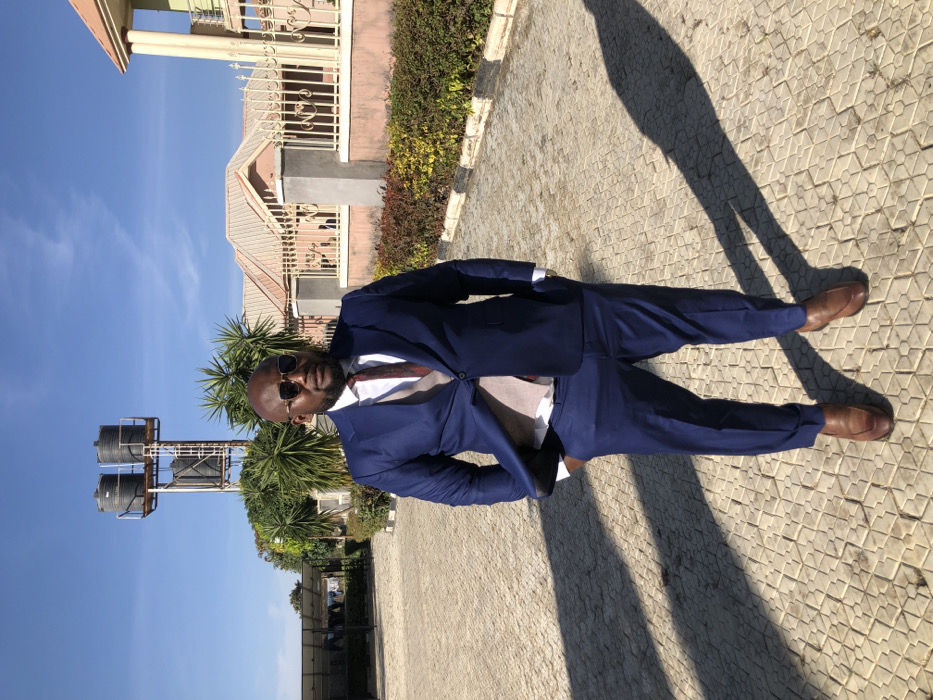 A man in a navy suit, light shirt, patterned tie, and sunglasses stands on a paved surface. He is wearing brown leather shoes. Behind him, there's a building with a beige facade, ornamental railings, and a bush of greenery. To the left, there are two black water tanks on a metal structure and a palm tree. A long shadow stretches from the man across the patterned ground toward the top right of the image, which has a bright, clear blue sky.