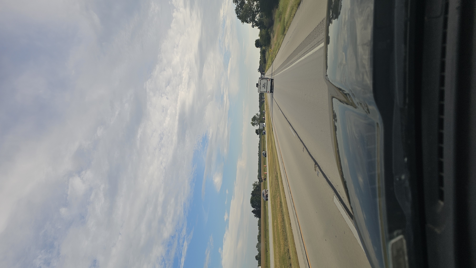 The image captures a view from a vehicle traveling on a highway, likely in or near Paxton, United States of America, under a partly cloudy sky. The perspective is from the driver's seat, showing the hood of the vehicle in the foreground. The highway stretches ahead, with multiple lanes marked by painted lines. On the left side of the highway, there are fields and trees bordering the road, with some scattered vehicles visible. To the right, more trees and vegetation line the roadside. A white recreational vehicle (RV) with "Jayco" branding is traveling in the same direction as the viewer's vehicle, ahead on the highway. The sky is a mix of blue sky and white, streaky clouds.