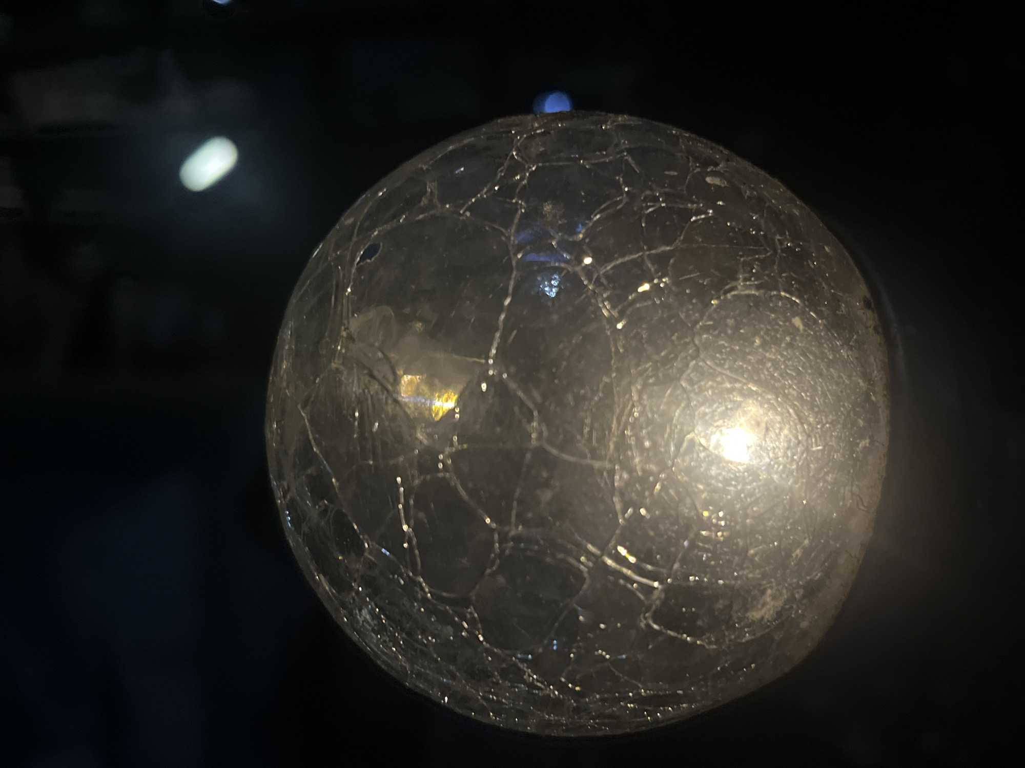 This media file displays a close-up, low-light shot of a cracked glass orb. The orb is illuminated from within, casting a warm glow that reflects off the fractured surface, creating a complex pattern of light and shadow. The cracks form an intricate network across the sphere, suggesting age or damage.
The background is dark and out of focus, with faint blurs of light that could be distant sources. One notable blur, seen in the upper left quadrant, is an elongated, soft white light. Another, more diffused light source is visible to the right of the orb, with a subtle gradient from light to dark. A small, intense blue light is visible on the top curve of the orb, possibly a reflection or an internal light. The overall impression is one of mystery and perhaps fragility due to the cracked surface and the isolated illumination.
No people are visible in the frame. The setting appears to be indoors or in a controlled environment where the orb is the primary subject. There are no discernible weather cues or specific location indicators, other than the provided context of Branipole, Bulgaria. The time of day is implied to be dark due to the lighting conditions. No text is visible in the image.