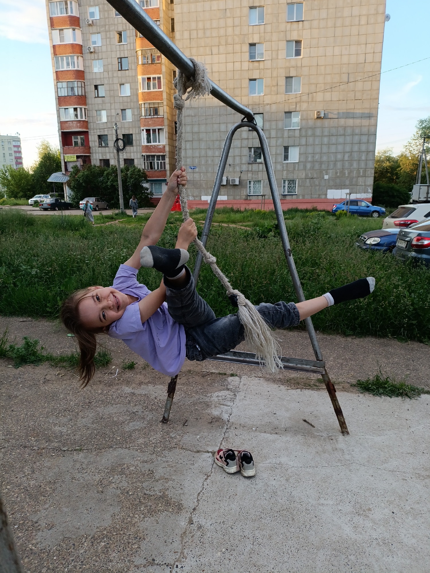 The image depicts an outdoor residential scene in the city of Chaikovsky, Russia. In the foreground, a young girl with light brown hair in a ponytail, wearing a light purple t-shirt, dark jeans, and black socks, hangs horizontally from a thick, frayed white rope. She is smiling directly at the camera. The rope is tied to a horizontal metal bar that forms part of a weathered, A-frame-like metal structure, possibly a ladder, with two vertical supports deeply set into the concrete ground. Directly beneath the girl, a pair of red and grey sneakers are placed neatly on the cracked, aggregated concrete surface.
The middle ground features a stretch of overgrown green grass and weeds. Beyond this, several vehicles are visible: a dark sedan and a light-colored sedan are parked on a dirt path to the left, while a bright blue sedan, a silver SUV, and another dark sedan are parked on a grassy area to the right.
In the background, two multi-story residential buildings dominate the skyline. The building on the left has a light-colored facade with numerous windows, red-brick balconies, and external air conditioning units. A yellow and white sign is partially visible on one of its lower balconies. Several figures, possibly adults, are walking on paths near this building. The building on the right is a tall, plain grey concrete panel structure with many windows and a few air conditioning units. The sky above is light blue with sparse clouds. Trees and bushes are visible behind the buildings, indicating a green, urban environment.