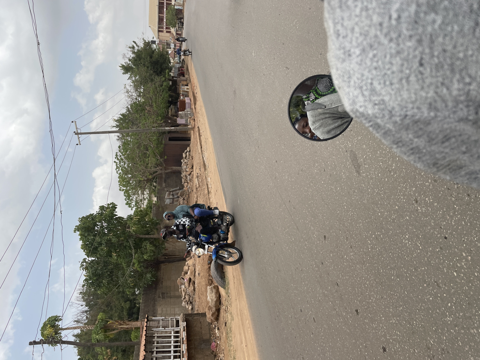 The image shows a street scene in Narayi, Nigeria. A motorcycle with three riders travels along the right side of the road. The road is paved and has some patches of dirt and debris. A shoulder and then a dirt and rock median are to the left of the road. A building and foliage is behind the dirt and rock median. Power poles are above the foliage. Buildings are in the background. A motorcycle’s side mirror with the reflection of a person is in the right of the image. A grey fabric is out of focus between the viewer and the mirror.