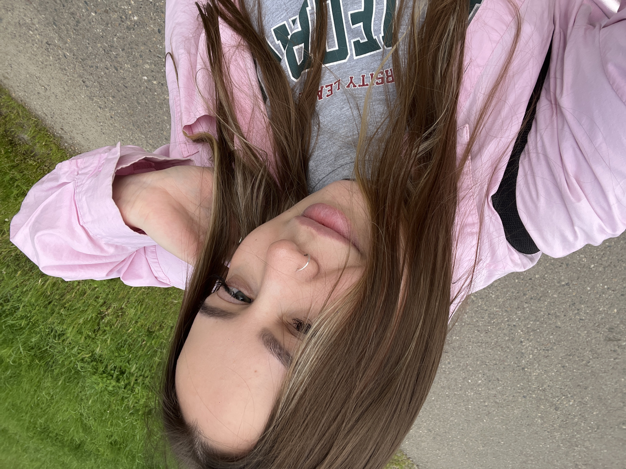 A young woman is lying on her side outdoors. She is wearing a pink jacket over a gray t-shirt with a dark green and brown graphic. She has long brown hair with some blonde highlights. She has a nose piercing on one side of her nose. The ground beneath her is grass and pavement. The image does not provide sufficient detail to ascertain the location in Novopokrovskaya, Russia.