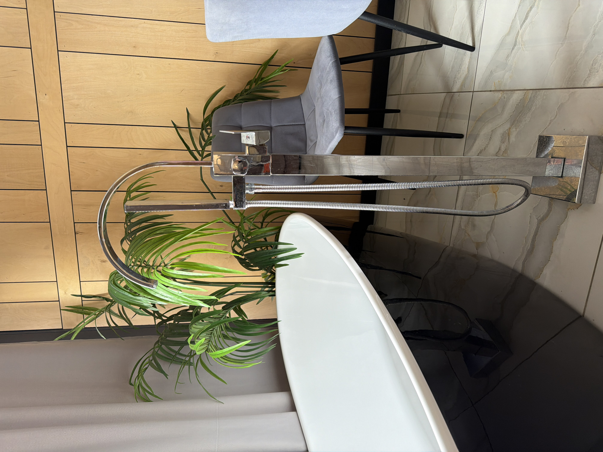 A freestanding bathtub faucet with a handheld shower attachment is positioned in the foreground of a bathroom. The faucet is made of chrome and features a curved spout. Behind the faucet, a lush green potted plant is visible. To the right, a grey upholstered chair with black legs is present, with another chair partially visible to its right. The background consists of a wall with vertical wooden panels and black trim. The floor is tiled with a marble-like pattern. The scene appears to be indoors, in a modern bathroom setting. There is no indication of the specific location within the city of Семилуки, Russia.