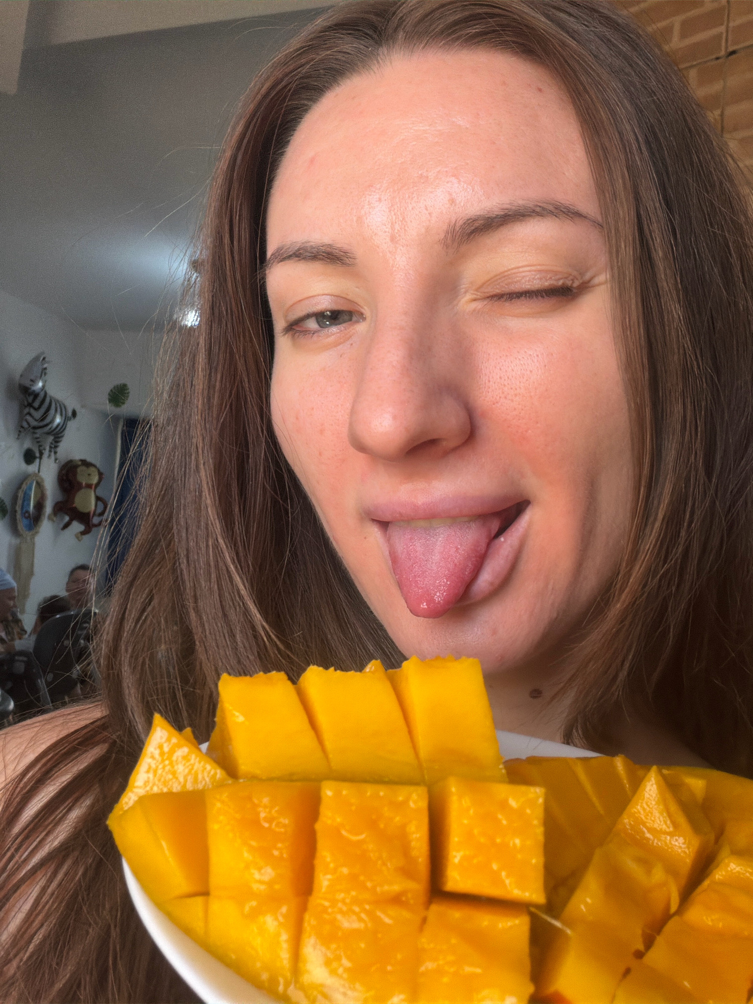 A young woman with brown hair is shown from the chest up, winking and sticking her tongue out. She is holding a white plate of sliced mangoes in front of her. In the background, there are some children's decorations, including a zebra balloon and a monkey figure. The woman appears to be indoors, possibly in a home or resort in Sharm El Sheikh, Egypt, though this cannot be definitively confirmed by the image alone.