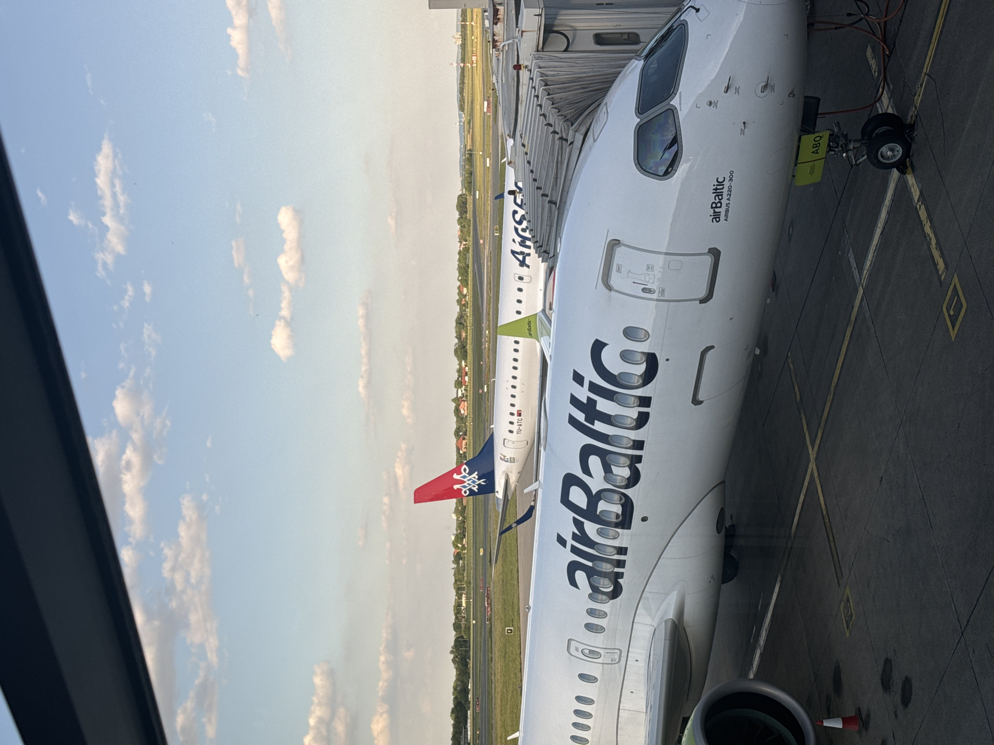 The image shows an AirBaltic Airbus A220-300 parked at an airport gate, likely at Belgrade Nikola Tesla Airport in Surcin Urban Municipality, Serbia, given the other planes visible. The AirBaltic plane is connected to a jet bridge. In the background, another aircraft, possibly an Air Serbia plane with a distinctive red and blue tail fin, is parked. The sky is visible in the background, with scattered clouds. The ground is paved with tarmac markings. The perspective is from inside the terminal looking out towards the parked aircraft.
