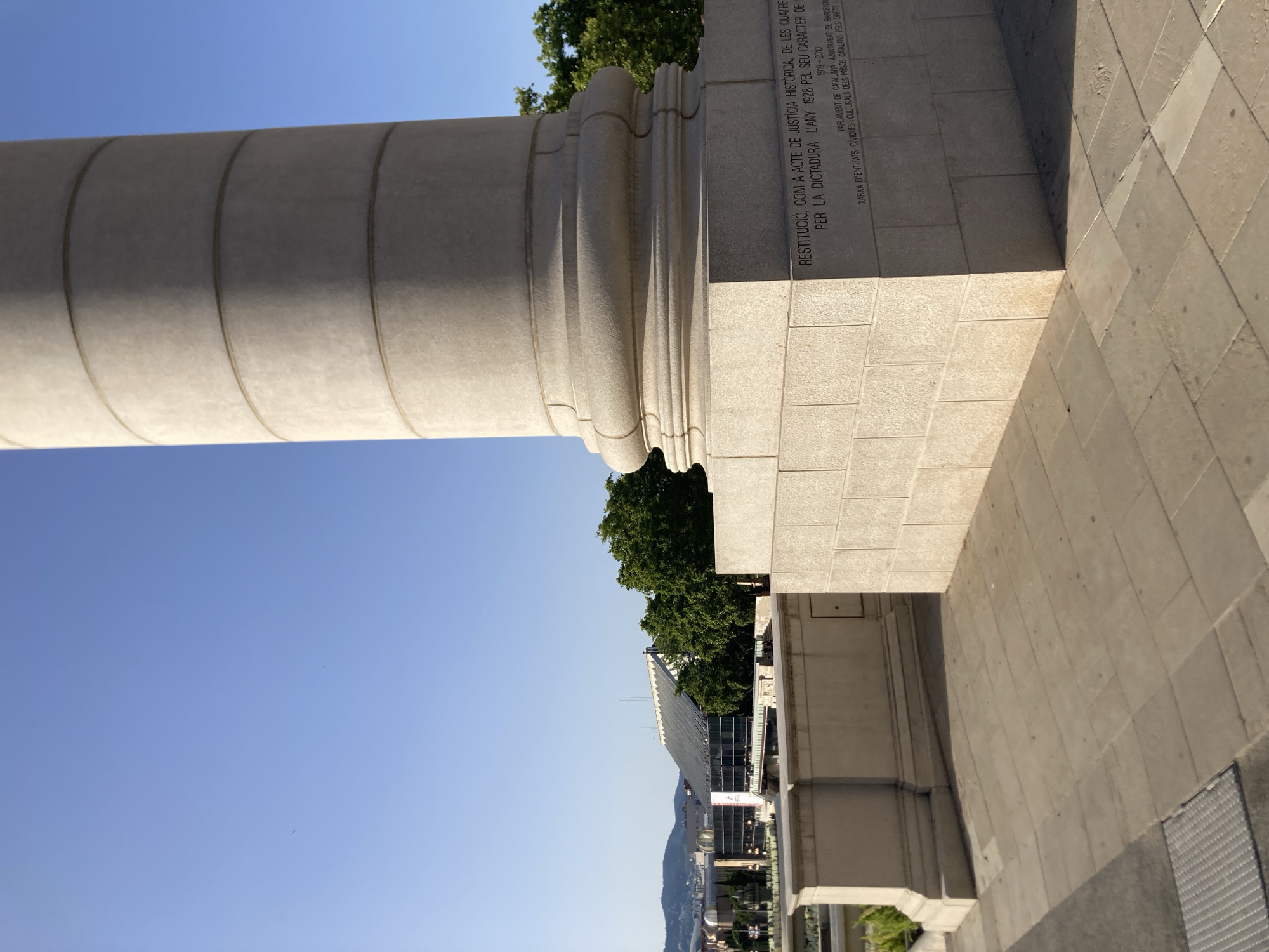 This is a close-up, eye-level outdoor photograph of a large stone column, part of a monument in Barcelona, Spain. The column is made of light-colored stone with horizontal bands. The base of the column is inscribed with text. The inscription reads: "RESTITUCIÓ, COM A ACTE DE JUSTÍCIA HISTÒRICA, DE LES QUATRE COLUMNES DE PUIG I CADAFALCH, ENDERrocades el 1924 PER LA DICTADURA. L'ANY 1928 PEL SEU CARÀCTER DE SIMBOL NACIONAL IMPULSAT PER LA DICTADURA 1919 - 2010 PARLAMENT DE CATALUNYA AJUNTAMENT DE BARCELONA XORXA D'ENTITATS CÍVIQUES I CULTURALS DELS PAÏSOS CATALANS PELS DRETS I LES LLIBERTATS". This translates to: "Restoration, as an act of historical justice, of the four columns by Puig i Cadafalch, demolished in 1924 due to the dictatorship. In 1928 for their character as a national symbol promoted by the dictatorship 1919 - 2010 Parliament of Catalonia, City Council of Barcelona, Network of Civic and Cultural Entities of the Catalan Countries for Rights and Freedoms".
The monument appears to be located near a public square or plaza, with a paved walkway in the foreground. In the background, a modern building with a glass facade and a large, sloping roof is visible, along with trees and distant hills. The sky is clear and blue. The lighting suggests it is daytime, with the sun casting shadows. No people are visible in the image.