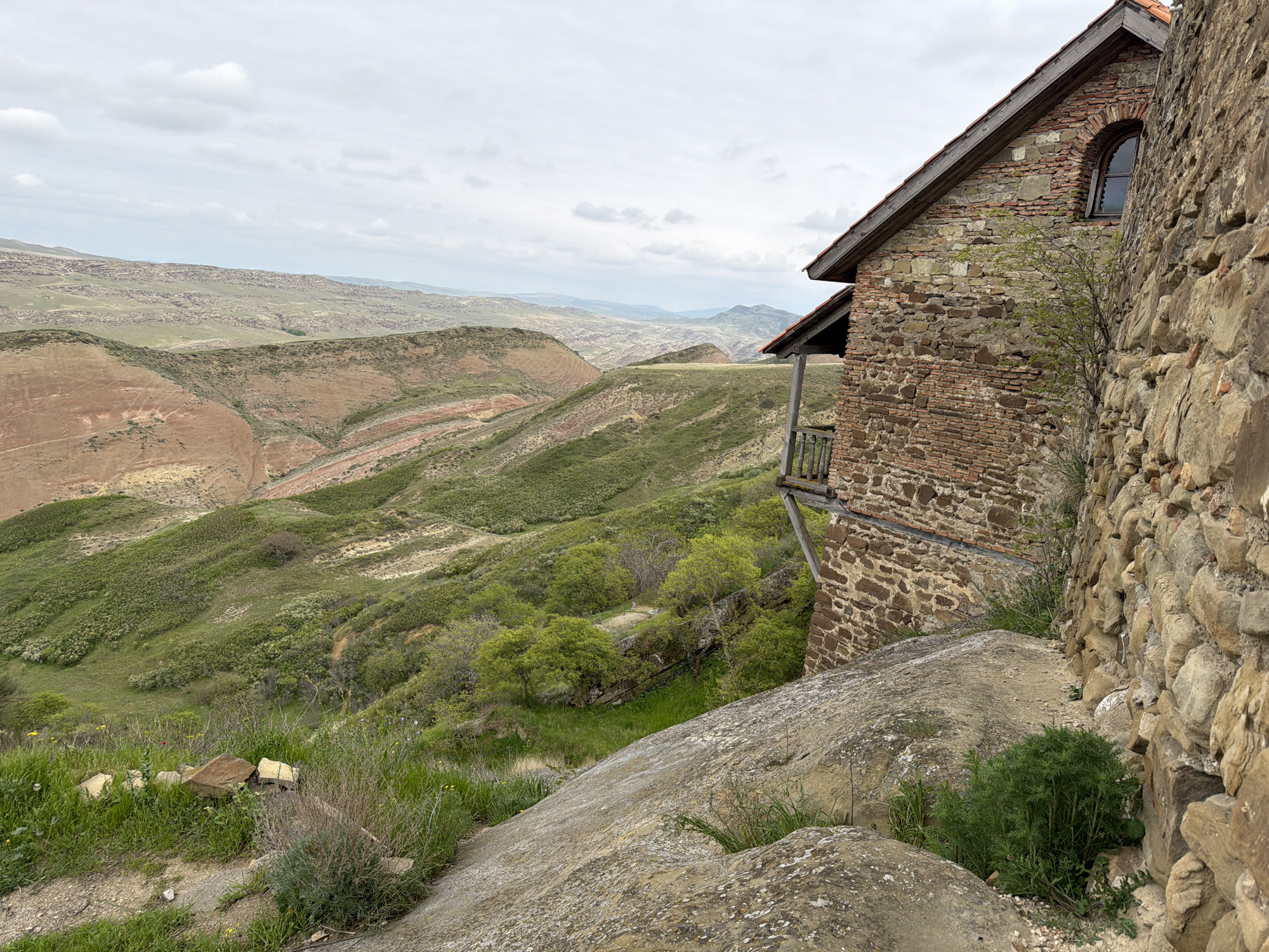The image presents a serene, remote landscape dominated by an ancient-looking building on the right side. This structure, possibly part of a monastery or historical complex, is constructed from a mix of robust stone blocks and reddish-brown bricks, indicating age and local building traditions. A small, arched window with a dark pane is visible high up on its side, beneath a roof lined with reddish tiles and supported by wooden beams. A simple wooden balcony with a railing projects from a lower section of the building, offering a vantage point over the expansive scenery.
The immediate foreground features large, weathered rocks and scattered green vegetation, including patches of grass and small bushes, suggesting a rugged, natural environment. The ground slopes sharply away from the building.
Beyond this, a vast, undulating hilly landscape unfolds under an overcast sky. The hills are characterized by a striking interplay of colors and textures: large areas are covered in lush green vegetation, while other sections reveal dramatic geological formations of exposed, reddish-brown earth, often displaying layered patterns. The distant hills appear softer and hazier, blending into the pale, cloudy sky. The overall impression is one of profound natural beauty and historical isolation, typical of the Udabno region in Georgia. The time of day appears to be daytime, under a uniformly gray, overcast sky. No human activity or visible text is present.
