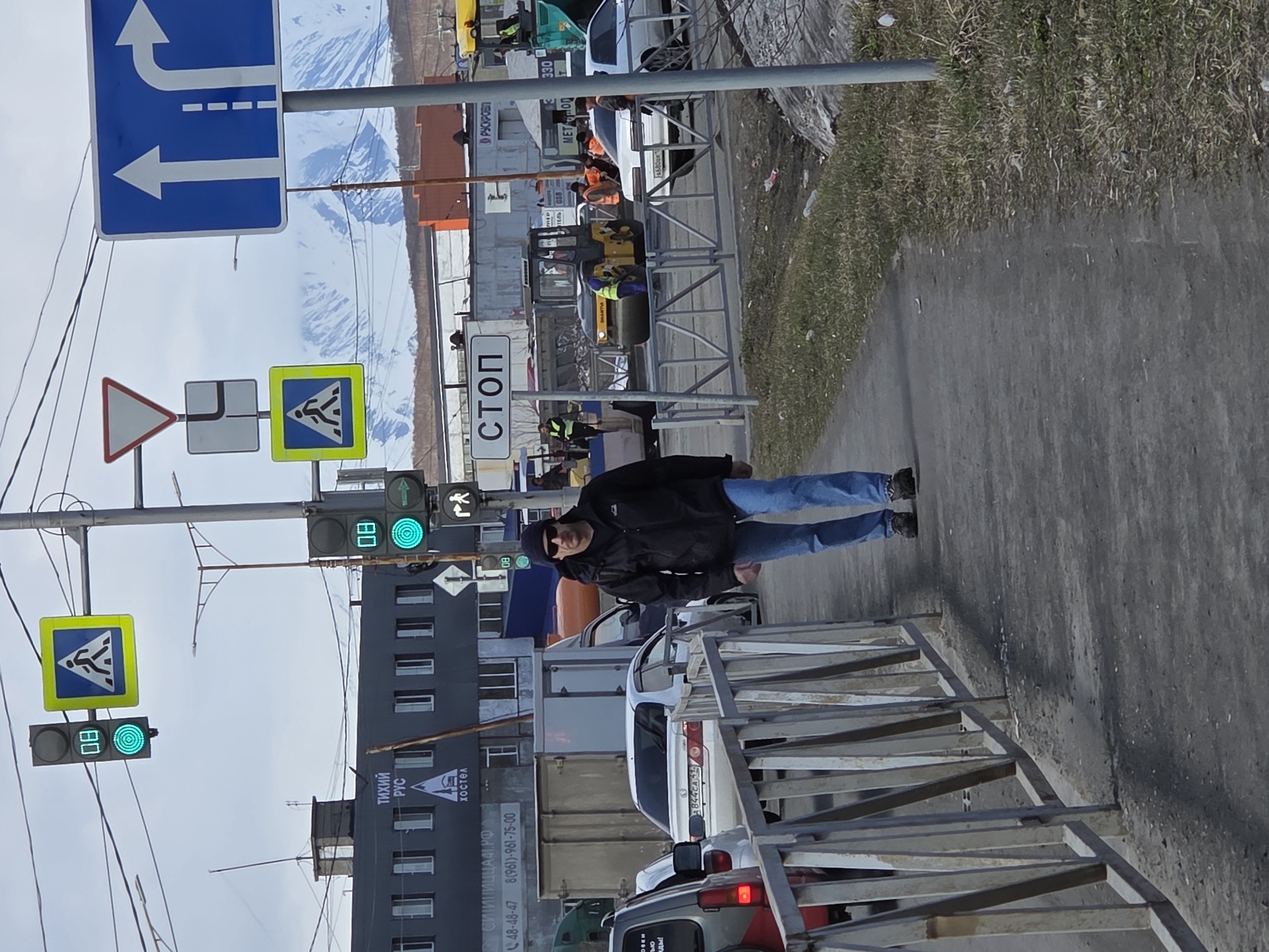 Here is a description of the image:
**Scene Overview:**
The image shows a street scene in a location identifiable as Unknown, Unknown, based on text on a building. The view includes elements of urban infrastructure like road construction, traffic signals, traffic signs, parked vehicles, and pedestrian walkways.
**People:**
One man is visible walking on the sidewalk. He is wearing jeans, a dark jacket, and what appears to be a dark beanie or cap.
**Objects:**
- Traffic lights are green for pedestrians and vehicles.
- Traffic signs are visible, including ones indicating a pedestrian crossing, direction of travel, a yield sign, and a stop sign.
- There are vehicles (cars and what seems to be a work truck) stopped or parked along the road.
- Construction barriers are present in the foreground.
- Buildings are visible in the background, along with mountain scenery further in the distance.
- There's signage visible on one of the buildings with text including "СИТИПИЦЦА", "ТИХИЙ РУС", and telephone numbers.
- Construction equipment is also visible.
**Actions and Interactions:**
- A man is walking on the sidewalk.
- Traffic is stopped, likely due to the signal and/or construction.
- Construction work appears to be in progress.
**Environment:**
- The setting is urban, with a street, sidewalk, and buildings.
- The sky is overcast.
- The presence of mountains indicates a geographical context.
- There is dry grass along the sidewalk.
**Location:** Unknown, Unknown.