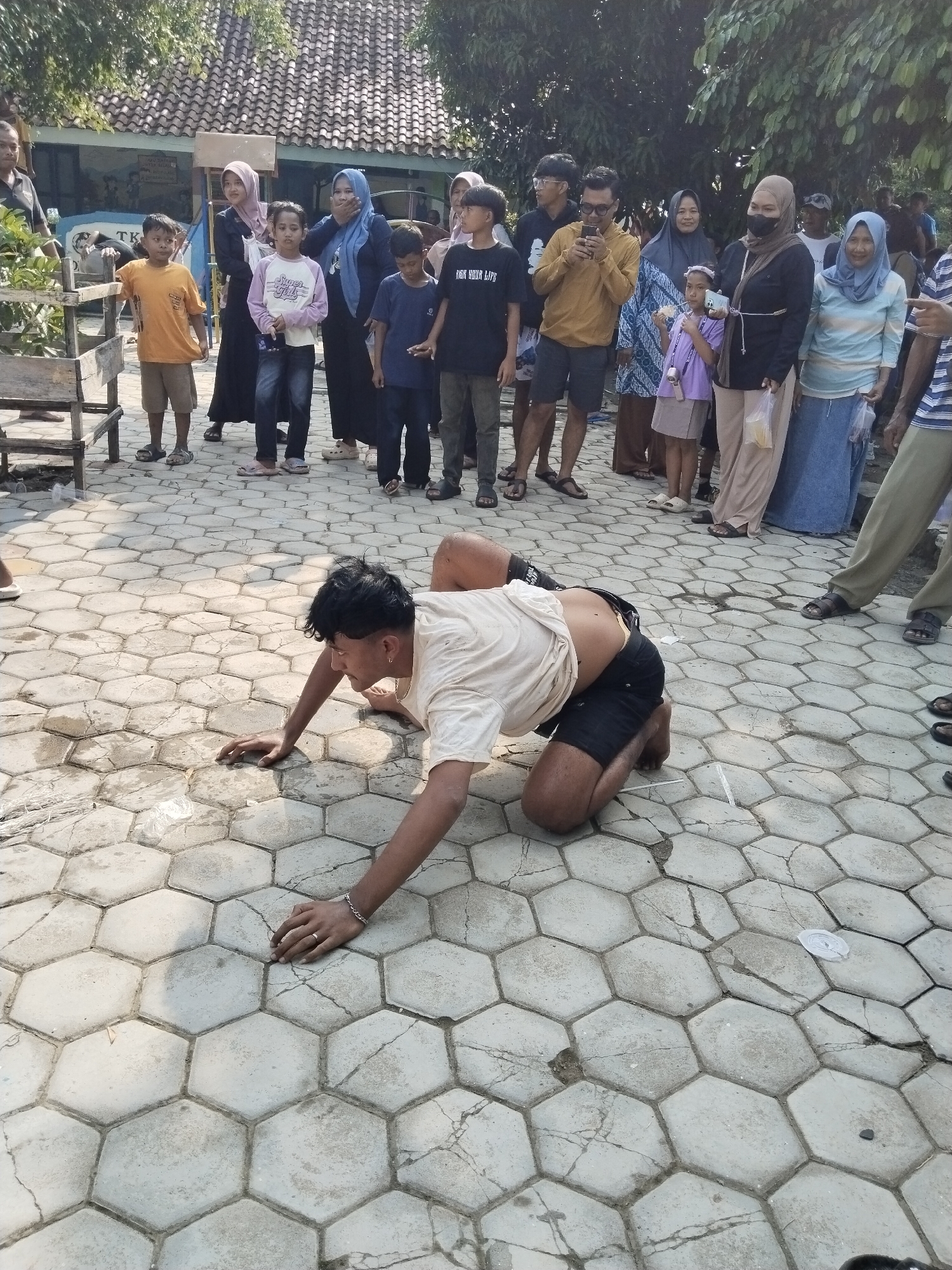The image, taken in Karangmalang, Indonesia, shows a man crawling on a hexagon-tiled surface. He wears a light-colored shirt and dark shorts. A crowd of onlookers, including women in hijabs and children, stands behind him. A building is visible in the background. The scene appears to be outdoors in daylight.