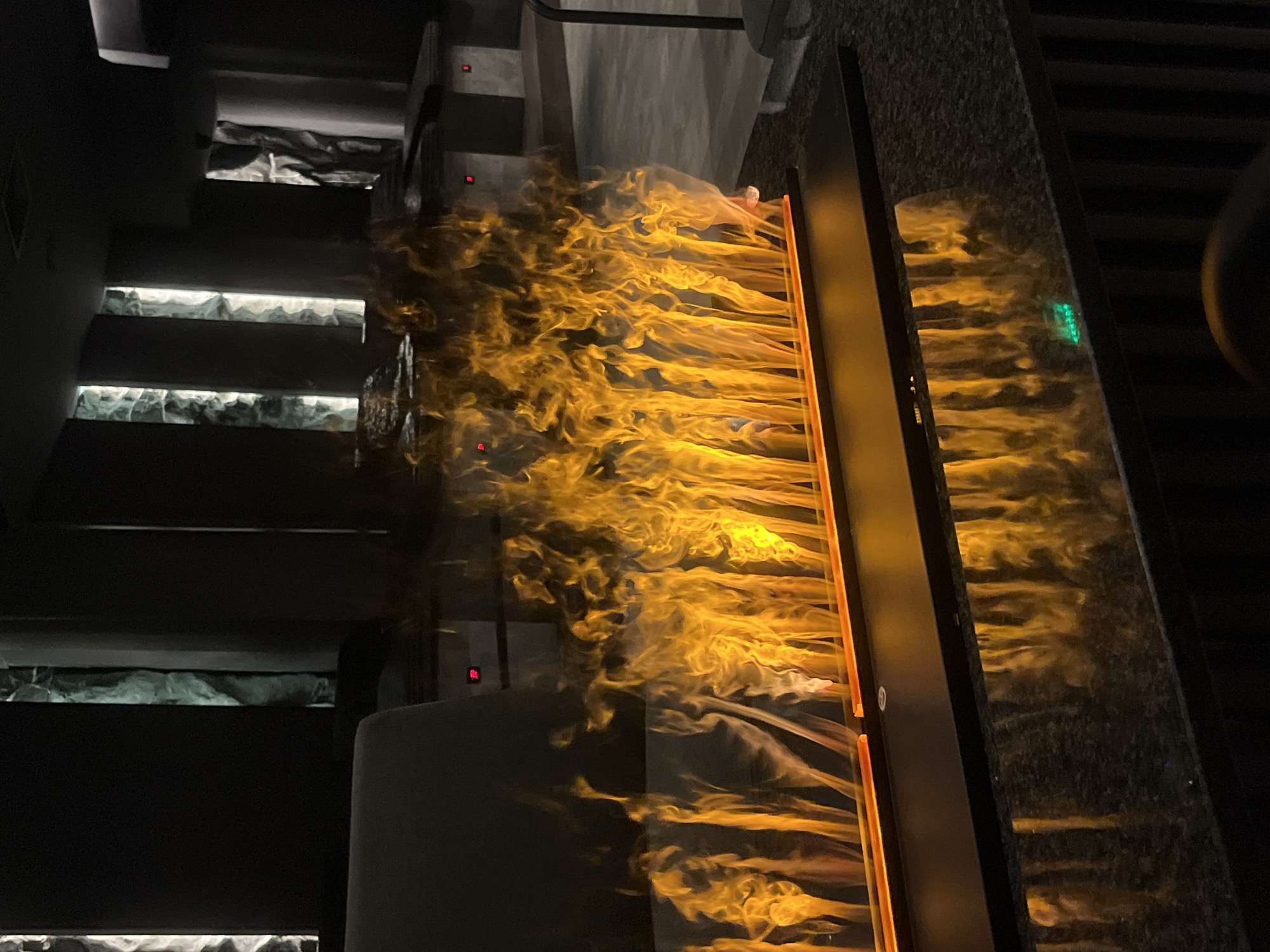 The image shows an interior space, likely a restaurant, in Sofia, Bulgaria. The scene is dark, with the primary light source being a stylized, flame-like wall feature that casts a golden glow throughout the space. A set of stairs with lit treads ascends on the left. The stairs appear to be concrete or a similar material, with a rough-textured surface. The wall feature is a long, vertical element with an internal light source creating the flame effect. The wall behind the feature has a dark, textured surface that reflects the light from the "flames." A portion of a dark chair is visible in the right foreground. The overall impression is of a modern, dimly lit, and possibly upscale establishment.