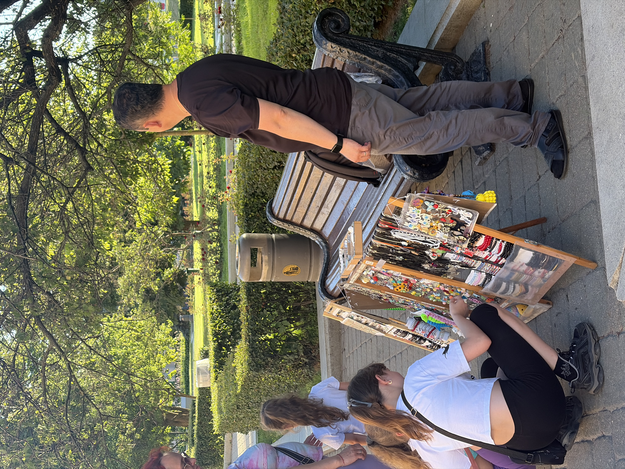 A man, a woman, and three young girls are in a park. The man is standing, looking away from the camera. The woman is partially visible on the left, wearing a multi-colored top. The three girls are looking at a display of jewelry and trinkets. One girl is squatting down, examining the items closely. The other two girls are standing behind her. The jewelry is displayed on wooden racks. In the background, there is a park bench, a trash can, and manicured bushes. The scene is illuminated by sunlight filtering through trees. The location is identified as Gorky Park, Minsk, Belarus.