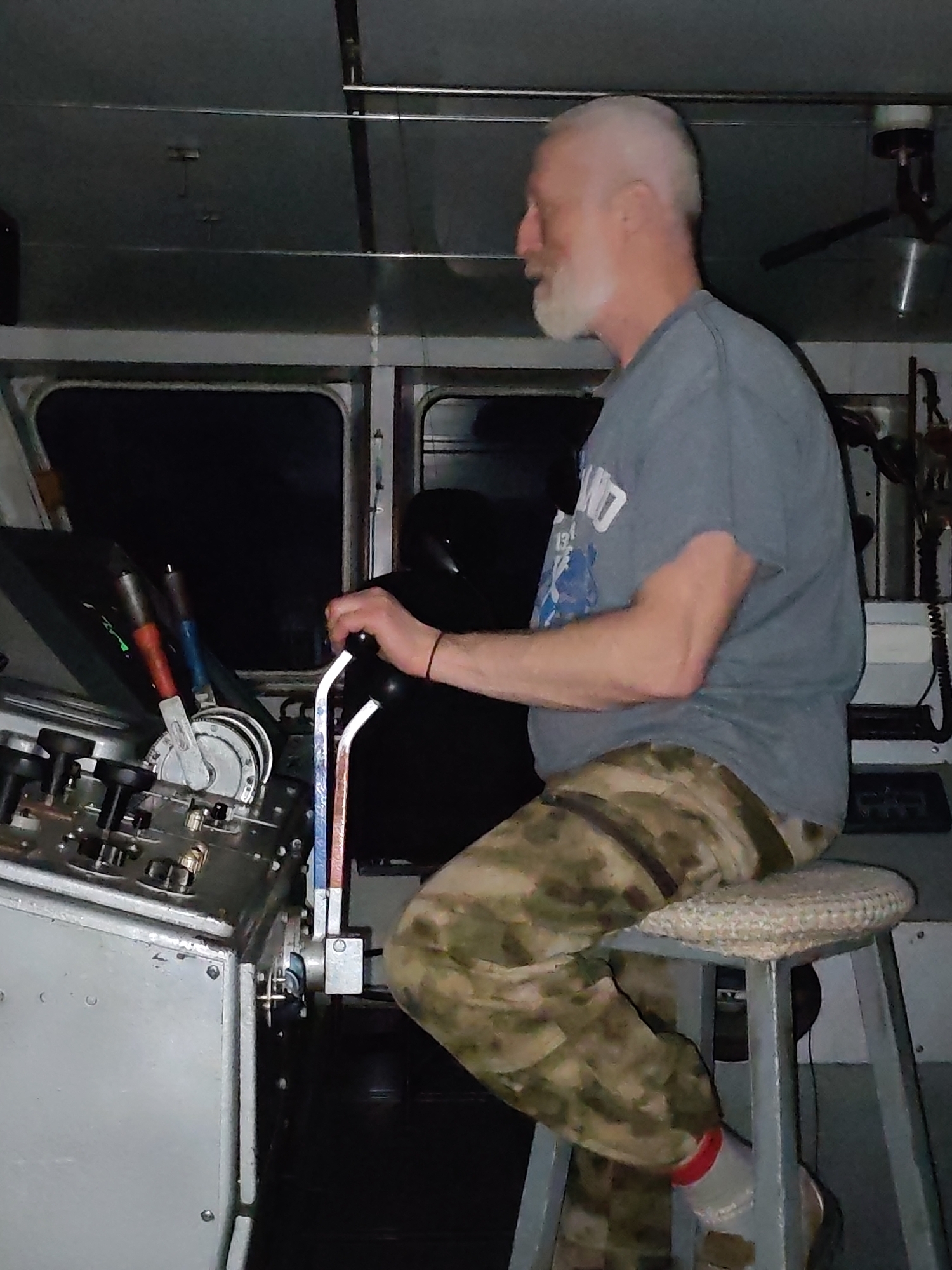 A man is seated on a stool inside a boat's control room. He has white hair and a beard, and is wearing a grey t-shirt and camouflage pants with a red ring around his right sock. He is holding a lever on a control panel that has several knobs and switches. The background is dark and shows windows and more equipment.