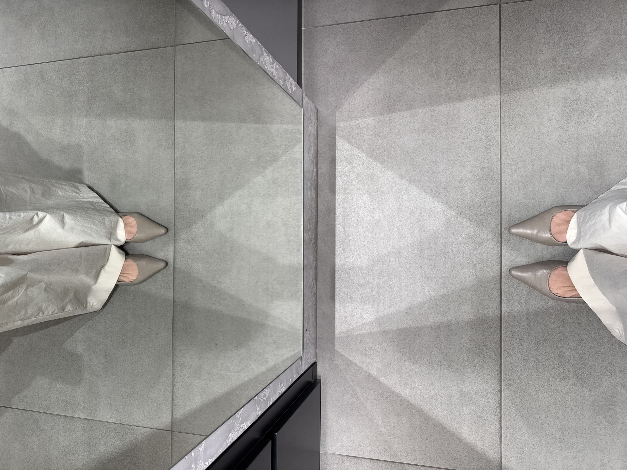The image shows a person standing on gray tiled flooring, wearing light-colored trousers and gray pointed-toe shoes. The person's reflection is visible in a mirror located to the left of the person. The mirror is set within a gray-toned frame. The flooring appears to be divided into large square tiles with dark grout lines. The scene is brightly lit, possibly indoors. There is no evidence to suggest the location is in Dubai, United Arab Emirates.