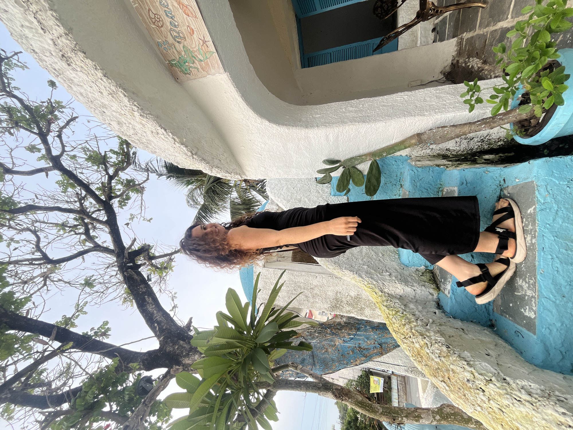 A woman stands outdoors, wearing a black dress and sandals, her curly brown hair framing her face. She is looking towards the camera with a slight smile. The scene appears to be a tropical or coastal area, given the presence of lush green foliage and palm trees. The architecture features brightly painted walls, primarily in white and a vibrant blue. A textured, white curved wall or structure dominates the foreground, with a painted mural featuring marine life visible at the top. The ground or steps are also painted in a striking blue. A potted plant with green leaves sits on a ledge. The overall impression is one of a sunny, perhaps warm, day in a visually colorful and relaxed environment. The image is oriented vertically.