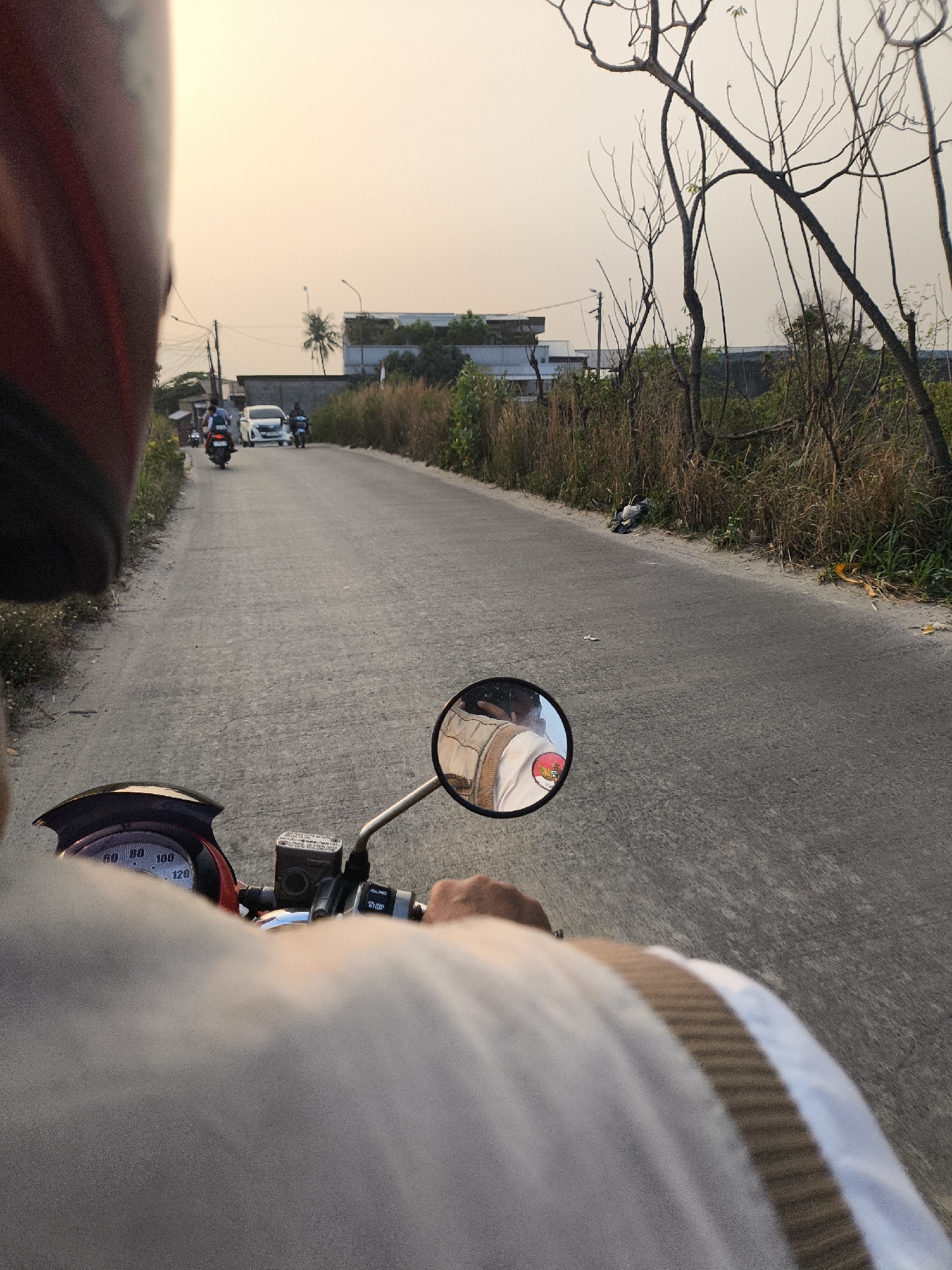 This is a first-person perspective photograph of someone riding a motorcycle on a paved road in Cilegon, Indonesia. The rider is wearing a red helmet and a light-colored jacket with brown stripes. The motorcycle's speedometer is visible, showing a speed of approximately 60 km/h. A circular rearview mirror on the motorcycle reflects the rider, who is wearing a white shirt with a circular emblem on the back, possibly a national symbol. In the distance, several other motorcycles and a white car are also on the road. The environment is rural, with dry vegetation and trees lining the side of the road. A building, possibly an industrial facility, is visible in the background under a hazy, pale sky, characteristic of sunset or sunrise. The road surface is asphalt.