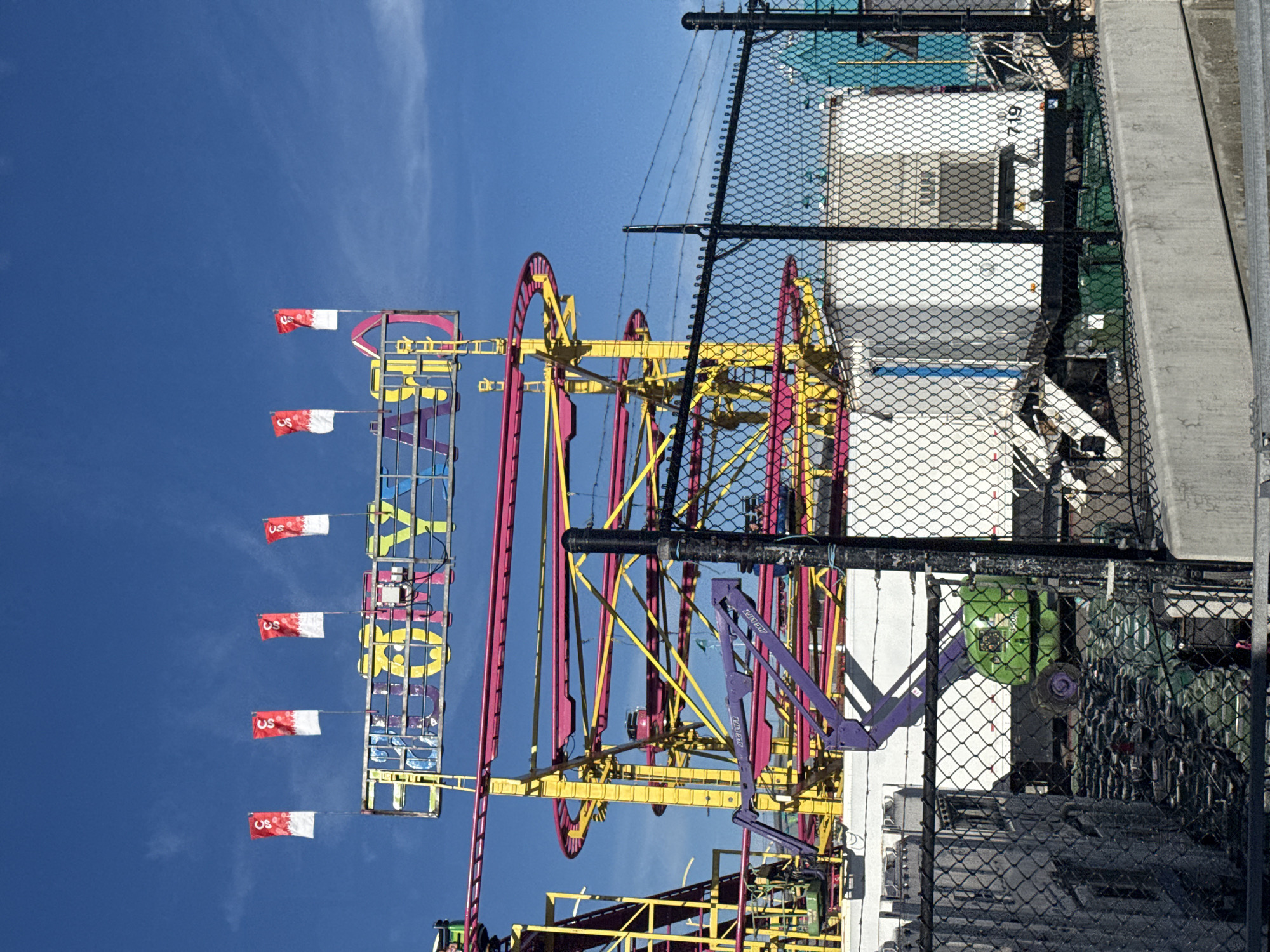 The image shows a bright, sunny day at a carnival or amusement park. A roller coaster with pink and yellow tracks dominates the scene. Above the roller coaster, a sign with colorful letters spells out "CRAZY AM." Four red and white flags with the letter 'S' are displayed on poles. In the foreground, a black chain-link fence is visible, with barbed wire running along the top. Behind the fence, there are white and blue trailers, a green maintenance vehicle with a purple lift, and stacks of what appear to be chairs or seating. The sky is a clear, vibrant blue with a few wispy clouds. There is no visible information within the image to suggest a specific location in Calgary, Canada.