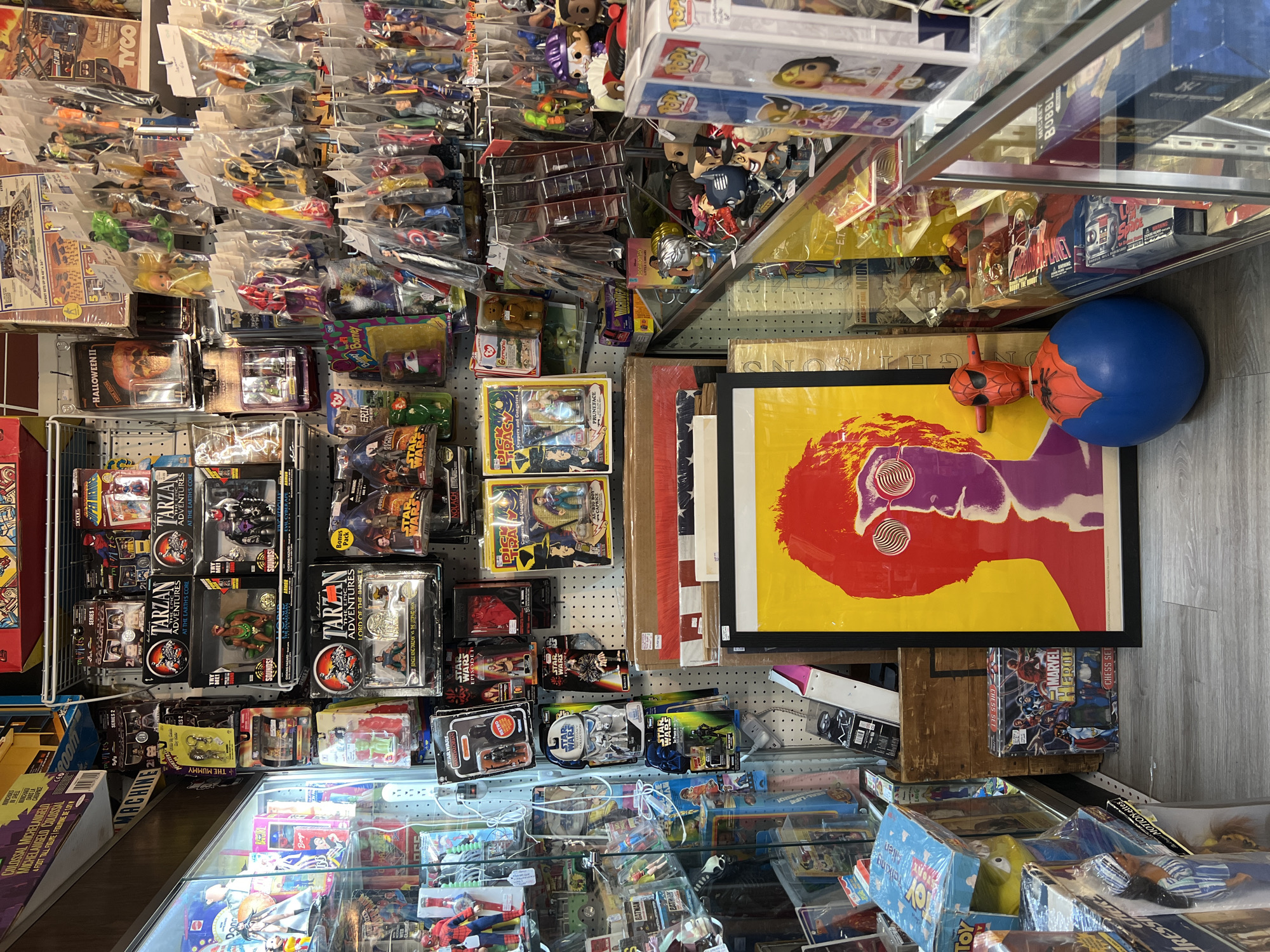 The image shows the cluttered interior of a store, possibly located in the City of Newburgh, United States of America, based on the camera's geotagging. The space is packed with merchandise, primarily toys and collectibles, arranged on shelves and in glass display cases. Items include action figures (e.g., "Tarzan," "Batman"), Funko Pop figures, vintage toys still in their packaging, and other memorabilia. A poster with a stylized graphic face is visible, along with a blue ball and a Spider-Man figurine. The overall impression is of a retail environment specializing in vintage or collectible items.
