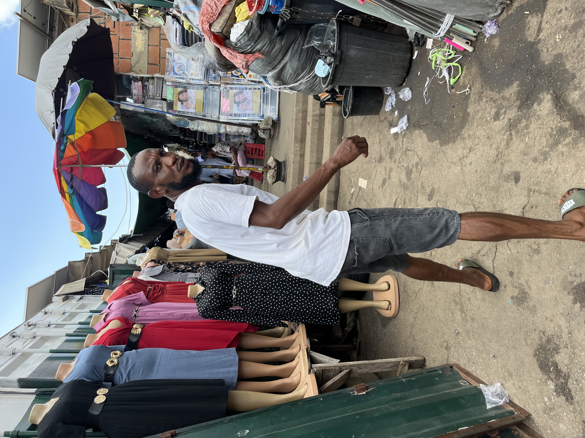 The image depicts an outdoor market scene, likely in Abuja, Nigeria. The foreground features a man standing, facing away from the camera, wearing a white t-shirt and grey shorts. His right arm is extended, and his hand is open, as if gesturing or reaching for something. To his left, a row of mannequins display various dresses in different colors, including black with white polka dots, red, pink, and blue. Some of the mannequins are adorned with belts.
Behind the man, the market unfolds. A large, open umbrella with multiple colorful panels is visible. Various stalls and makeshift structures are present, with goods stacked and displayed. Bags of what appear to be trash or merchandise are piled up. In the background, there are glimpses of what might be election-related posters with images of people. The ground is paved with a rough, concrete-like surface, showing signs of wear and some scattered debris. A large black bin is also visible.
The lighting suggests it is daytime, possibly mid-morning or afternoon, with shadows cast on the ground. The weather appears to be fair, with a clear sky visible in the upper left portion of the image. The overall atmosphere of the scene is that of a busy, bustling marketplace. The man's expression is not clearly visible, but the context of the market suggests an environment of commerce and daily life.