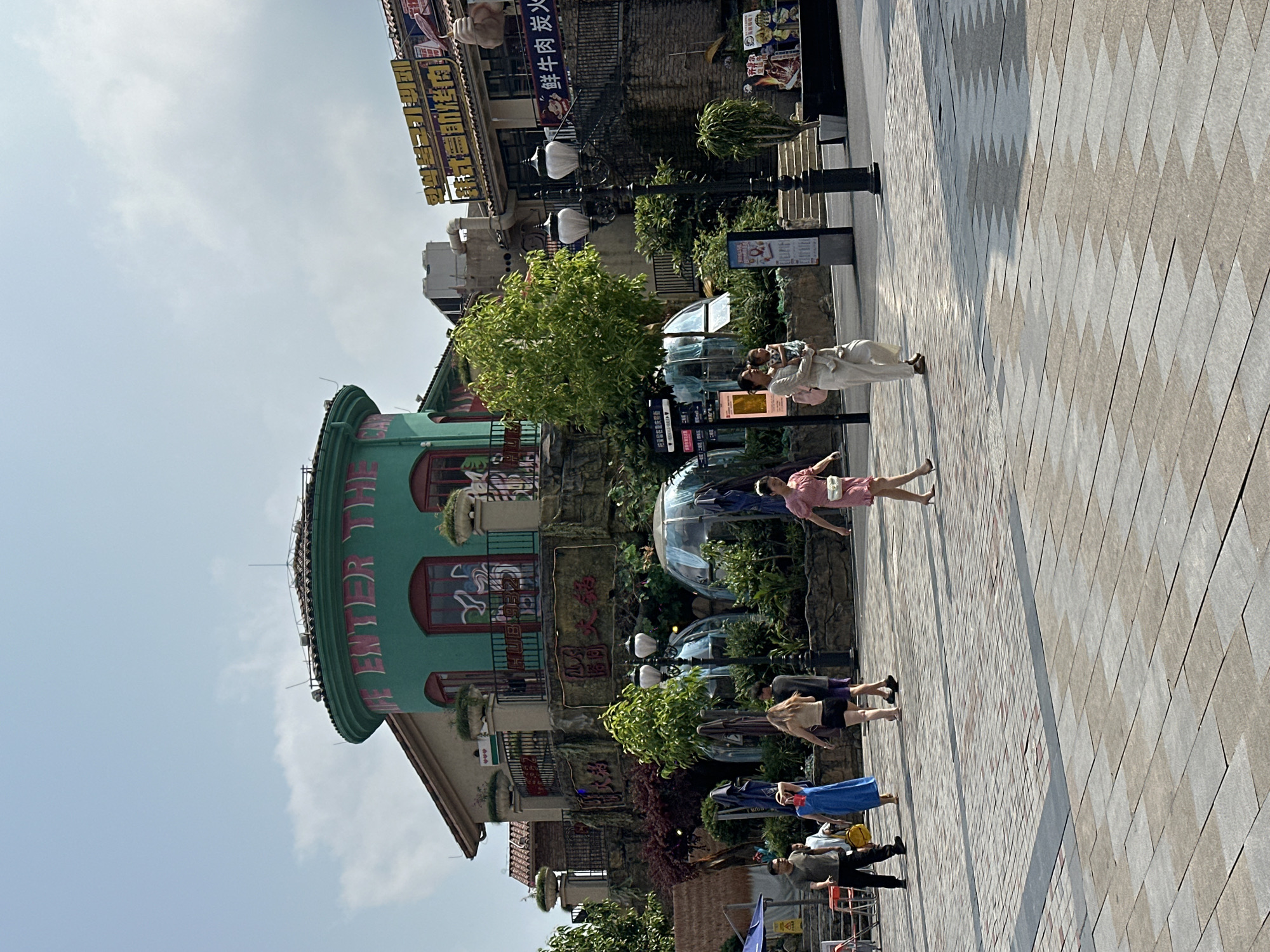 The image shows a street scene in the Chongqing High-tech Zone, China. Several pedestrians are walking on a paved area, including a woman in a pink dress and a woman holding a baby. There is a distinctive green building with the words "ENTER THE" written on its side. There are also some trees, bushes, and streetlights in the background, as well as a building with Chinese writing.