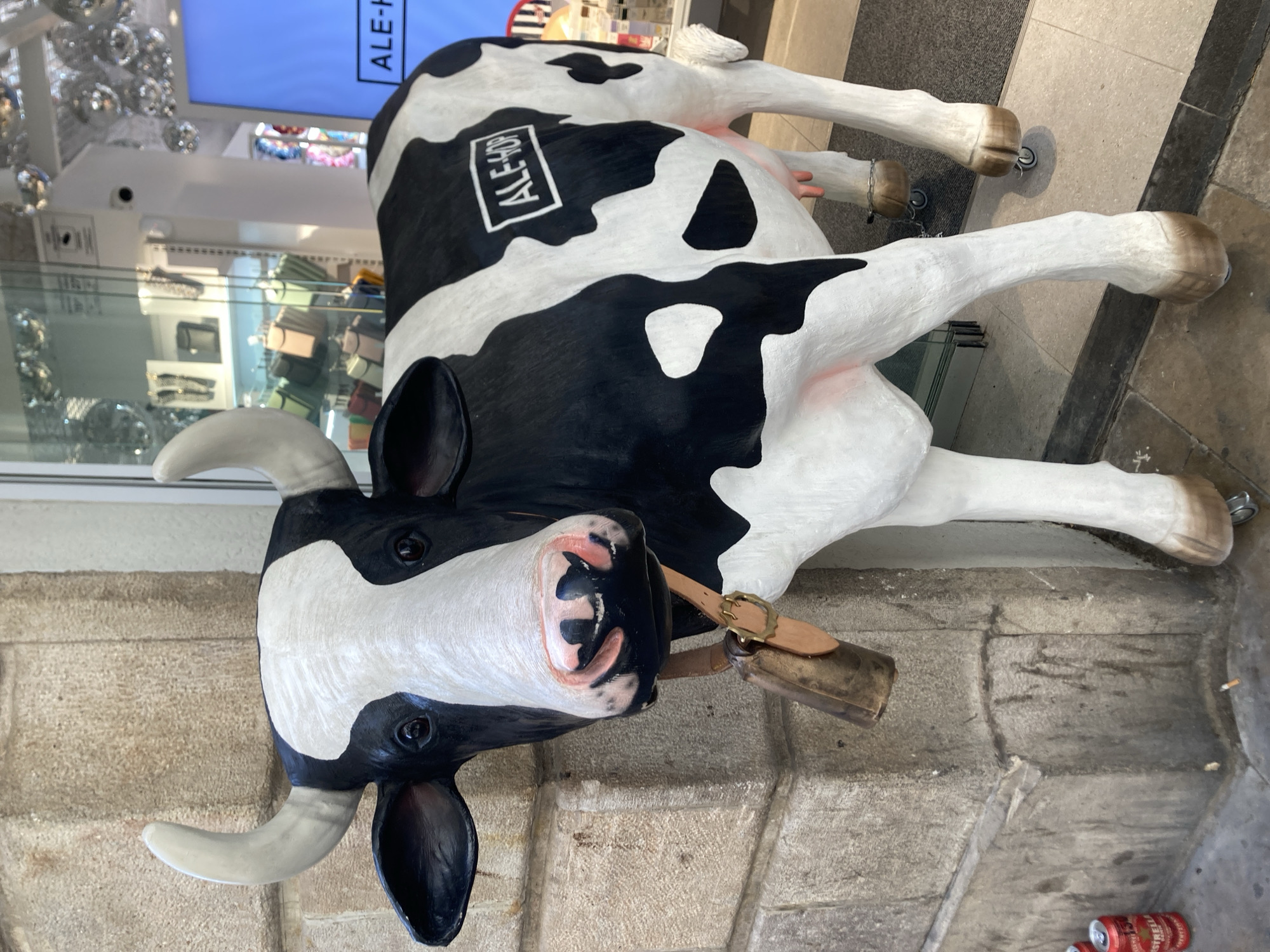 A life-sized, black and white cow sculpture is the main subject, positioned on its side. The cow has a visible bell around its neck, and a leather strap with a buckle. The sculpture's head is tilted, with its mouth open, revealing pink gums and black interior. One horn is pointing upwards, the other downwards. The cow has a white rectangle on its side with black text.
Behind the cow, a retail environment is visible. There are shelves displaying various items, possibly bags or accessories, in pastel colors. Above these shelves, a large screen displays text, and further back, there are disco balls reflecting light. The floor appears to be paved with stone tiles. The overall setting suggests an outdoor area, likely a commercial street or plaza, in Barcelona, Spain, given the context provided. The lighting suggests it is daytime, and no specific weather conditions are apparent. There are no people in the image.
Visible text includes "ALE-HOP" on the cow's side and on the screen in the background. There are also some red and silver cans in the lower right corner of the image.