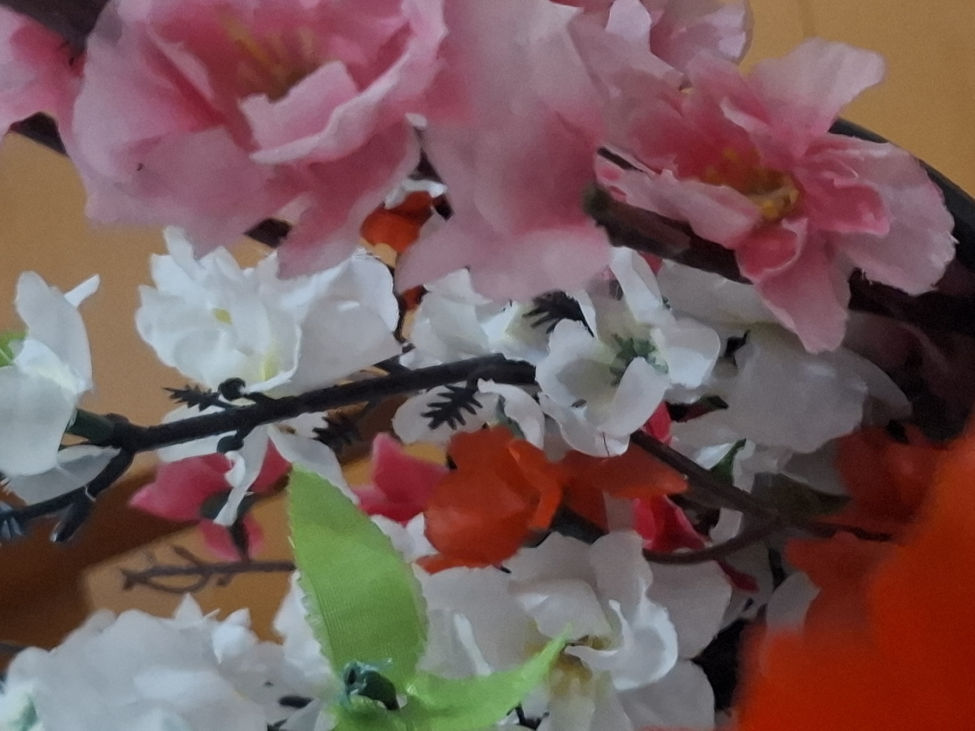 The image displays a close-up view of a bouquet of artificial flowers, featuring various colors including white, pink, orange, and green. The flowers are arranged on dark stems. A blurred red object occupies the lower left portion of the image, likely part of the container or another floral element. The background is a warm, out-of-focus yellow or tan. The image does not contain any identifiable landmarks or elements that would allow for a determination of its location within Almaty, Kazakhstan.