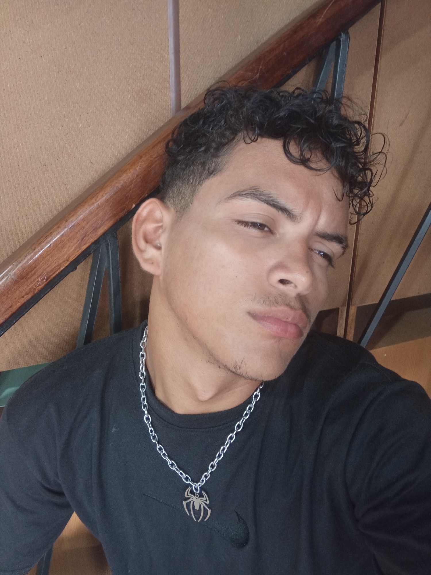 A young man with short, curly black hair and light brown skin is pictured from the chest up. He wears a black t-shirt and a silver chain necklace with a spider pendant. He is looking slightly off-camera with a furrowed brow and a faint mustache and beard. The background includes a wooden handrail with black metal supports and a beige paneled wall. The location in Juan Griego, Venezuela is not evident from the image.
