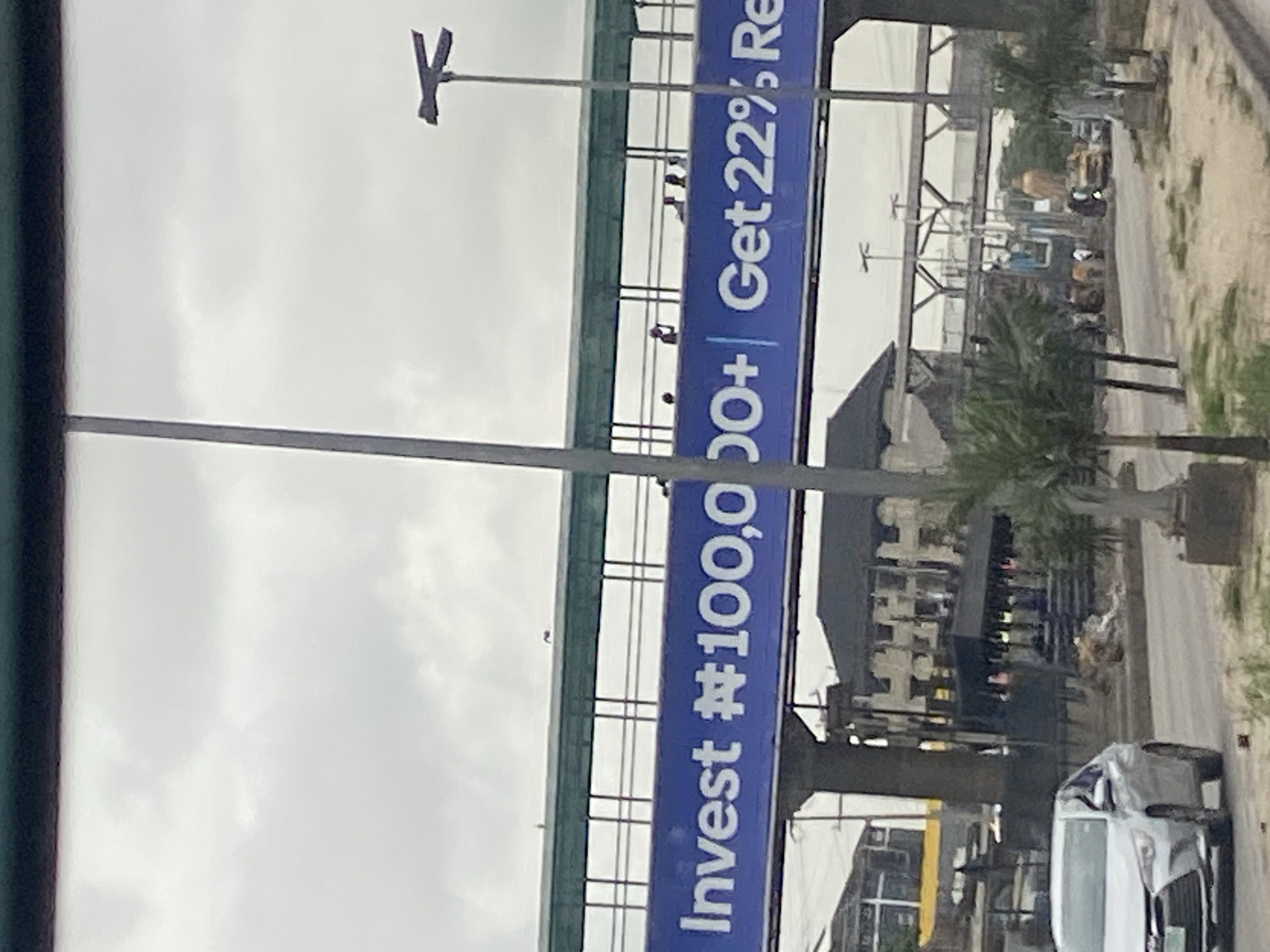 The image depicts a pedestrian bridge in Lagos, Nigeria, adorned with a prominent blue banner. The banner displays white text that reads, "Invest ₦100,000+ Get 22% Re." Several individuals are visible standing on top of the pedestrian bridge, some of whom appear to be silhouetted against the overcast sky. Below the bridge, a silver sedan is driving on a road. The surrounding environment includes buildings, trees, and utility poles, consistent with an urban setting in Lagos.