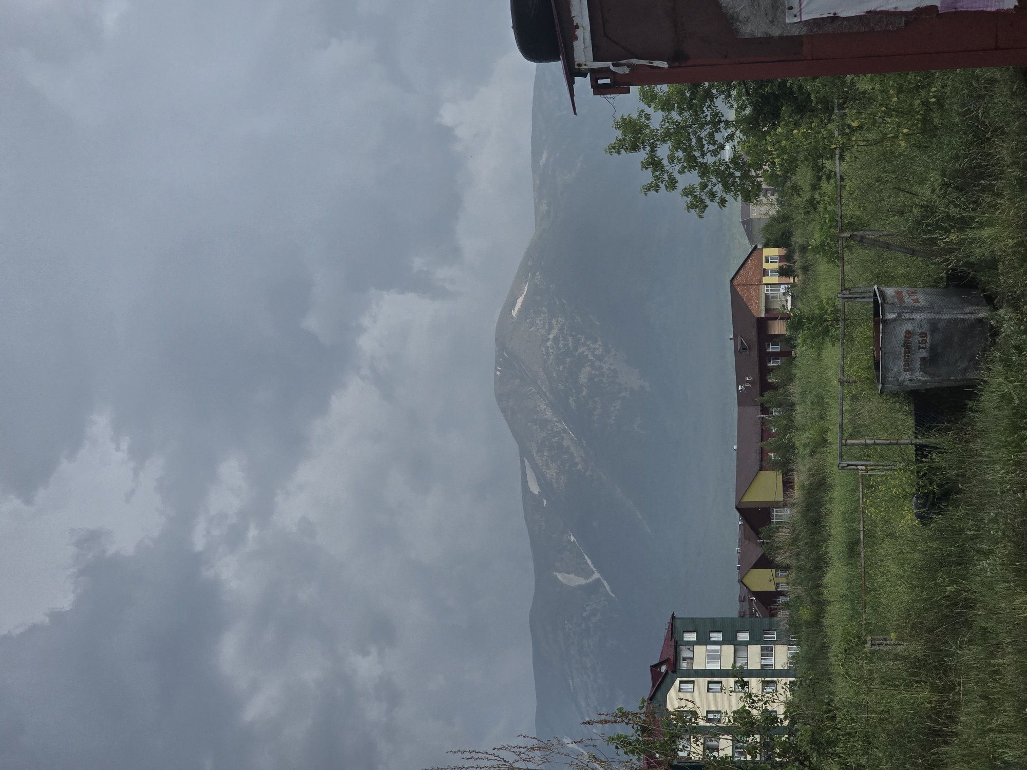 The image shows a cloudy sky over a mountainous landscape. In the foreground, there are several buildings, including a tall, light-colored apartment building with green accents, and several smaller, lower buildings with yellow and red facades. There is a lot of green grass and vegetation in front of the buildings, with a metal fence and a trash bin visible. To the far right, a portion of a dark-colored structure with a tire on top is visible. The scene appears to be a residential area near a mountain range. The location is identified as the city of Ridder, Kazakhstan.
