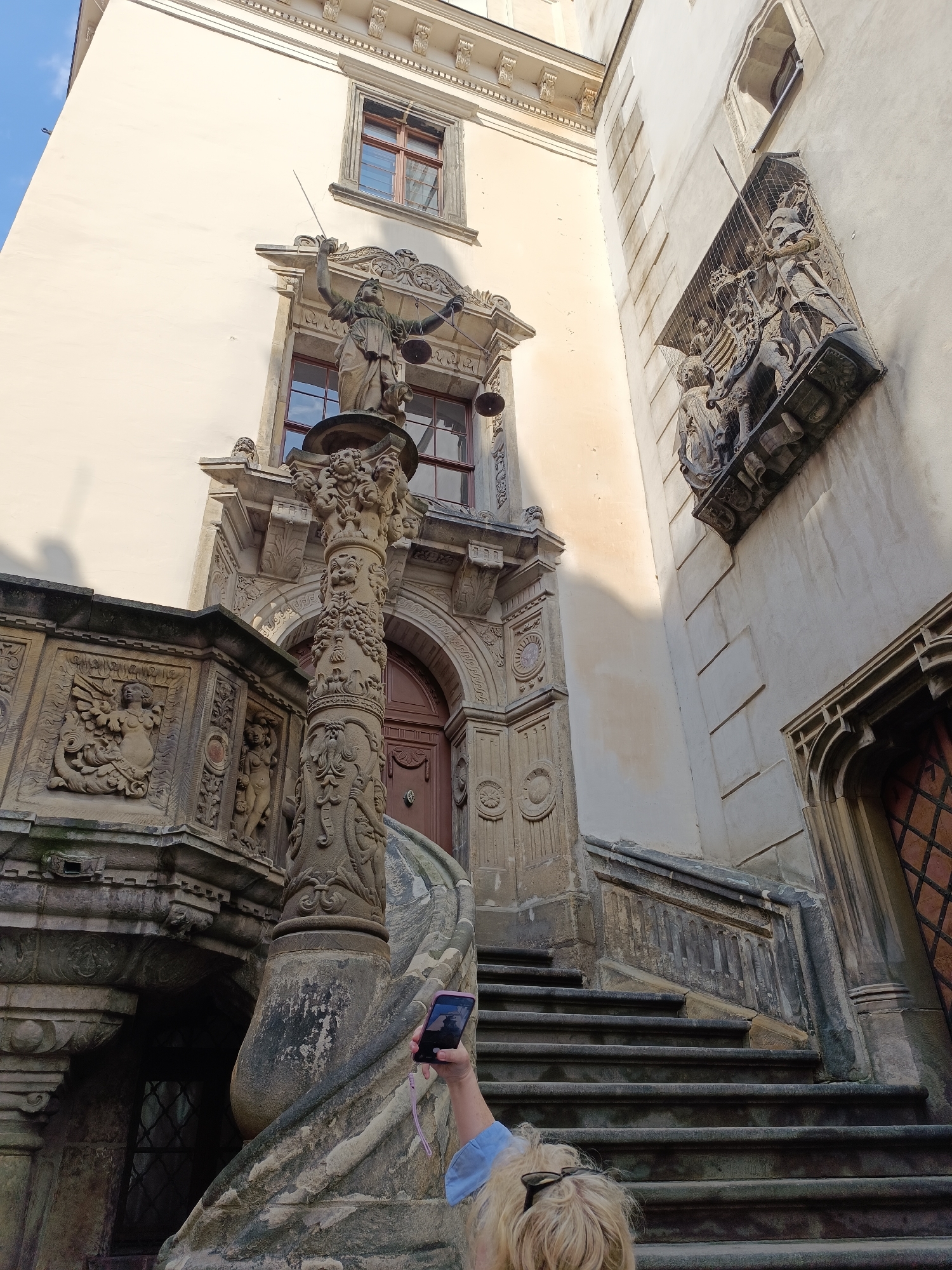 The image shows a stone staircase leading up to a building. The staircase is flanked by ornate stone carvings, including a statue of Justice holding scales. A person is visible at the bottom of the stairs, holding up a smartphone to take a picture of the statue. The building appears to be old and has a weathered facade. The sky is visible in the upper left corner. This scene is located at the Kaisertrutz in Görlitz, Germany, which is a historical fortification that now houses a museum.