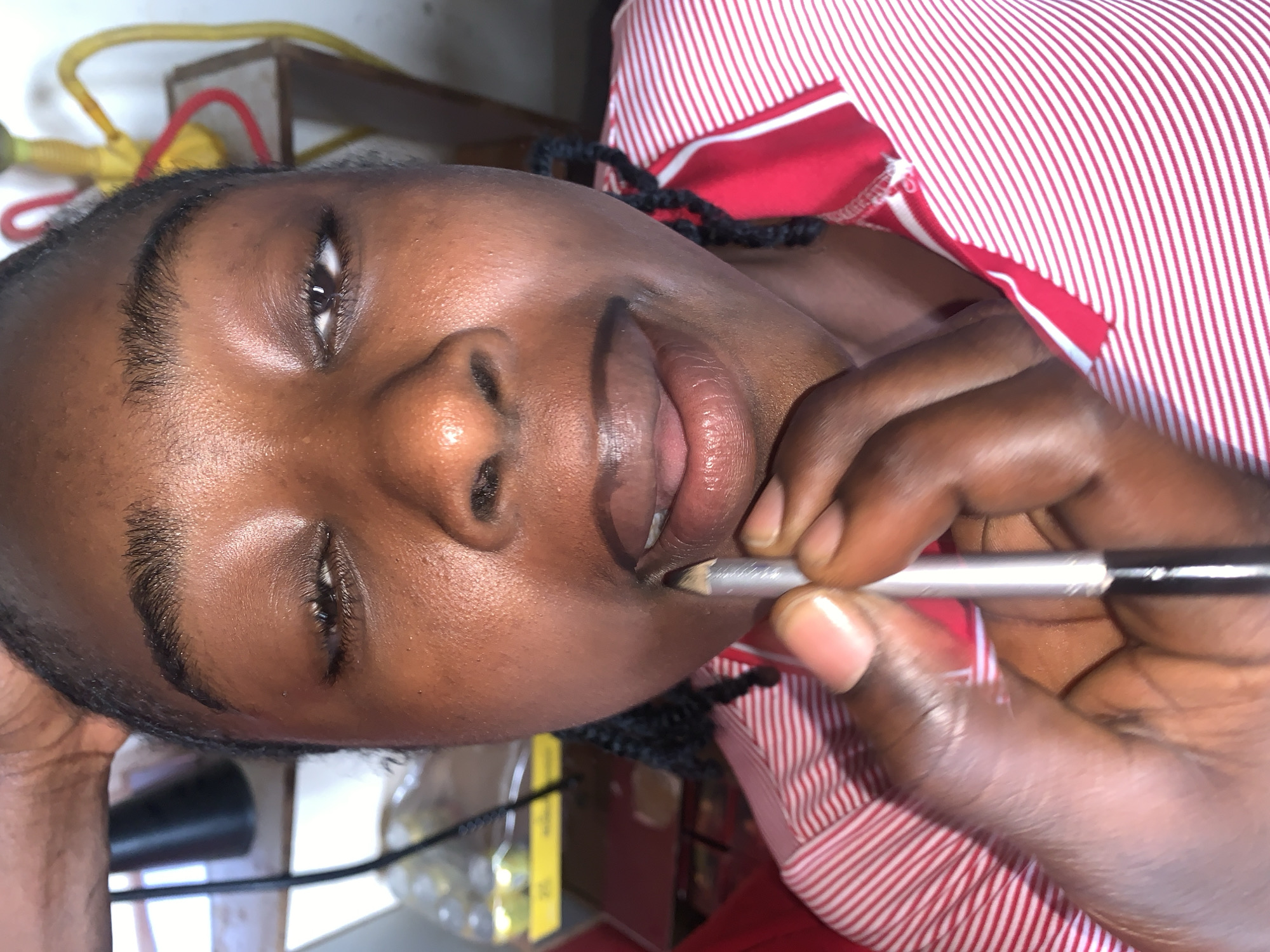 The media file displays a close-up, rotated portrait of a person, likely a young woman, with dark skin, engaged in applying makeup. She is seen from a slightly elevated angle, with her head positioned towards the left side of the frame. Her dark hair is styled in braids, partially visible near her neck and behind her head. She wears a red and white striped top, possibly a shirt or dress.
The primary activity is the application of a product to her lips. Her right hand, with visibly healthy nails, holds a silver-bodied makeup pencil with a dark tip. The pencil is positioned at the corner of her mouth, suggesting she is lining or filling her lips. Her lips are slightly parted, revealing some inner lip tissue.
Her facial expression conveys a sense of focus, accompanied by a subtle smirk or gentle smile. Her eyes are open, with the right eye (viewer's left) looking slightly upward. Her eyebrows are dark and well-defined. The skin on her face has a natural sheen, particularly on her forehead and cheeks.
The background is softly blurred and indistinct, showing various shades of light and dark. Hints of yellow and red wires or cords are visible, along with what appears to be a wooden surface or furniture. The setting appears to be indoors. No specific time of day or weather conditions can be determined from the image. No visible text is present. The described location context for this image is New Stadium, Nigeria.