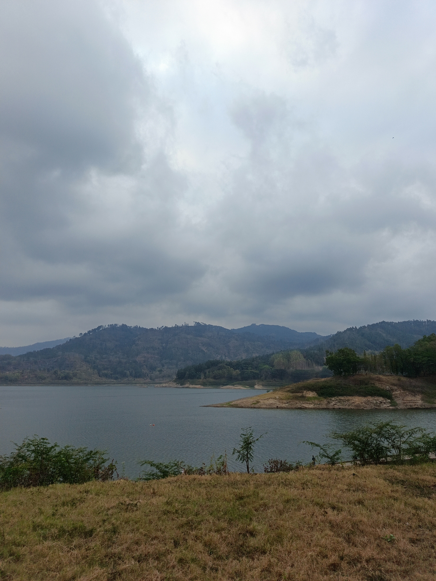The image depicts a wide, tranquil lake under a cloudy sky. Rolling hills covered in sparse vegetation form the backdrop, with a few trees dotting the landscape. The foreground is a grassy slope, dry and autumnal in appearance. On the lake, two small orange buoys are visible. In the middle ground, near the shore, a small boat with two orange markers is present. Two figures are visible on the right bank. This scene is located in Segawe, Indonesia.