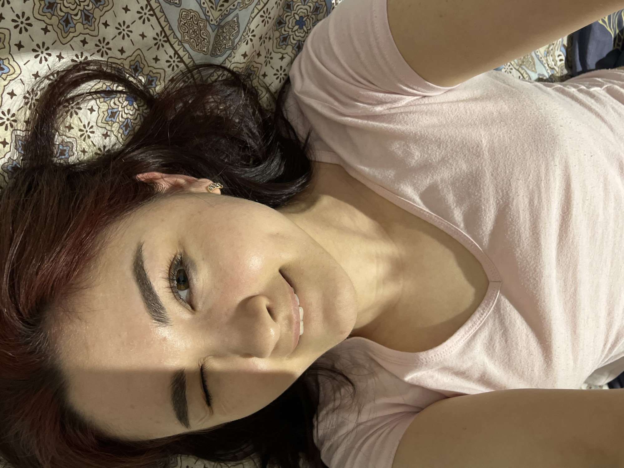 A woman with dark hair, partially dyed reddish-brown, is lying on a bed with patterned bedding. She wears a light pink v-neck t-shirt. She is looking directly at the camera with a slight smile, partially closing one eye. A shadow falls across her face. There is no information to indicate this photo was taken in Neftekamsk, Russia.