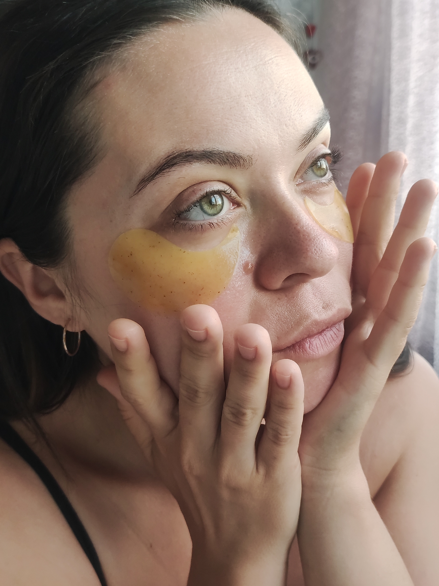 A woman is seen with gold eye patches under her eyes. Her hands are held up near her face. She has dark hair and green eyes. The scene appears to be indoors, possibly near a window. It is not possible to determine the specific location, including whether it is in Chaikovsky, Russia, from the image alone.