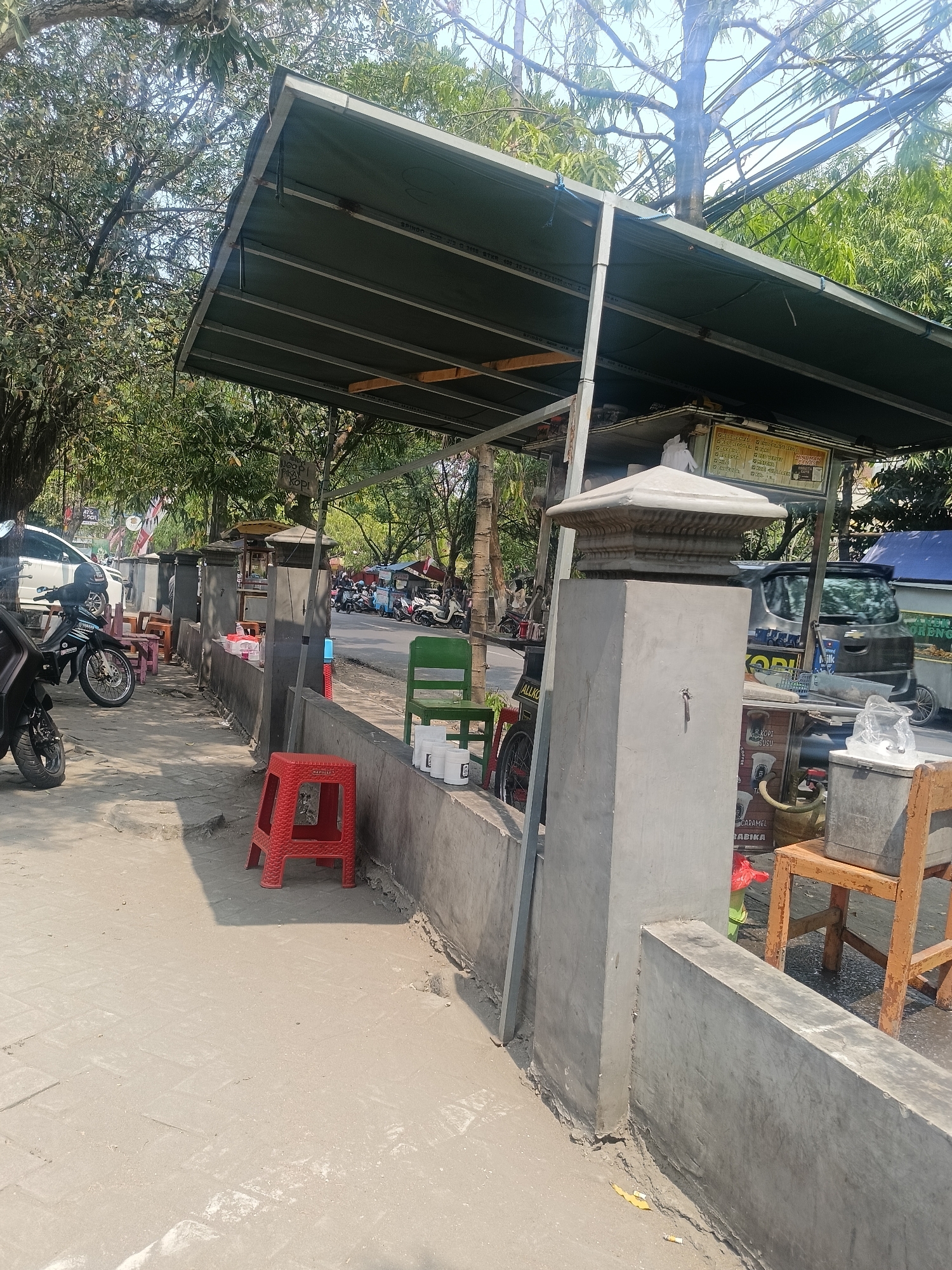 The image depicts a street scene in Kediri City, Indonesia, likely a roadside food stall area. A covered structure with a green awning extends over a walkway, providing shade. Several small, colorful plastic chairs and a red plastic stool are placed along a low concrete wall. In the background, motorcycles and cars are parked along the street, suggesting it's a busy thoroughfare. There are also a few food carts visible, with signage indicating beverages like coffee and milk. Trees and power lines are present overhead, contributing to the urban outdoor environment. The ground appears to be paved with concrete or stone. The overall impression is of a casual, open-air dining or resting area catering to passing traffic.
