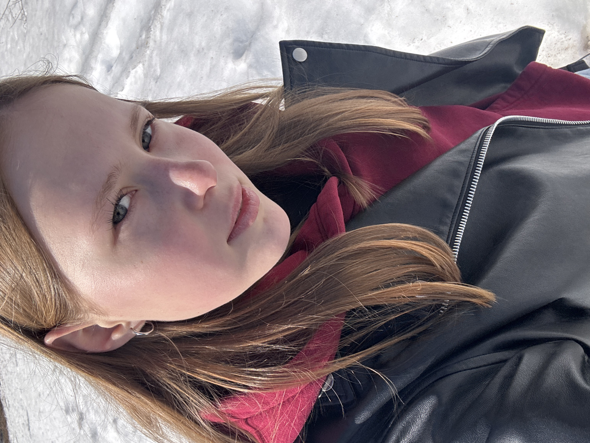 A young, light-skinned female lies on her side on snow-covered ground. She wears a maroon hooded sweatshirt under a black leather jacket. Her light brown hair is spread around her head. An earring is visible in her left ear. The photo appears to have been taken in Chaikovsky, Russia, though this detail is not directly observable within the frame.