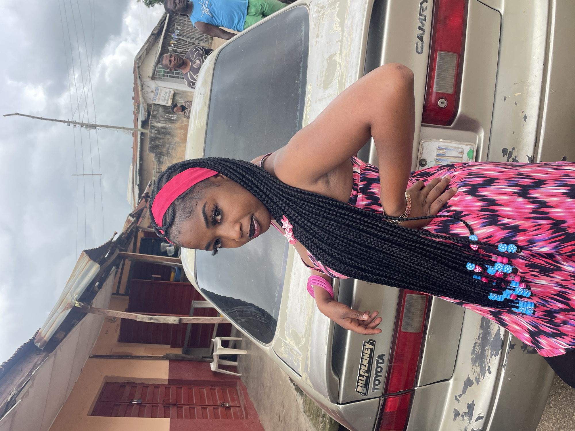 In Jos, Nigeria, a young woman with long, dark braided hair adorned with blue and pink beads stands leaning against the trunk of a light-colored Toyota Camry. She is wearing a pink and black patterned dress and a pink headband. Her left arm is bent at the elbow, resting on the car, with her hand on her hip. Her right hand is visible near her hip as well, with a gold bracelet. Behind her, two men are standing near the car, and in the background, there are buildings with corrugated metal roofs and some electrical poles. The sky is overcast.