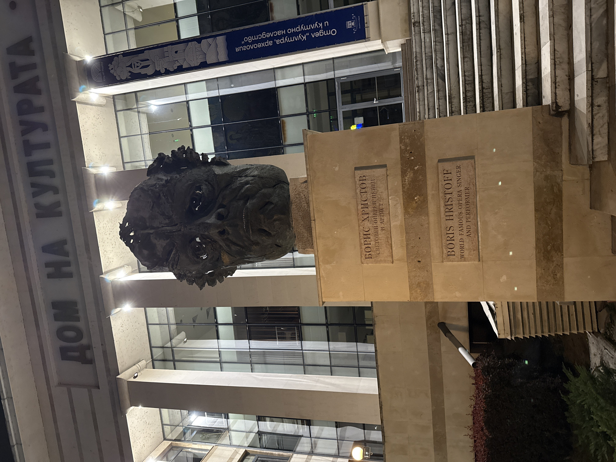 The image captures a bronze bust of a man, Boris Hristoff, positioned outdoors at night. The bust, featuring a rugged texture and a solemn expression, rests on a light-colored stone pedestal. Two plaques are visible on the pedestal. The upper plaque, in Cyrillic, reads "БОРИС ХРИСТОВ СВЕТОВЕН ОПЕРЕН ПЕВЕЦ И АРТИСТ" (BORIS HRISTOFF WORLD FAMOUS OPERA SINGER AND PERFORMER). The lower plaque provides the English translation: "BORIS HRISTOFF WORLD FAMOUS OPERA SINGER AND PERFORMER".
Behind the bust stands a large, modern building with a facade featuring glass panels and light-colored columns. On the left side of the building, vertical text in Cyrillic reads "ДОМ НА КУЛТУРАТА" (HOUSE OF CULTURE), identifying the structure as a cultural institution. High on the building's exterior, above the bust, a blue banner is displayed. The banner's inverted text reads "Отдел „Култура, археология и културно наследство“" (Department "Culture, Archaeology and Cultural Heritage"). A small blue and yellow flag, possibly indicating support for Ukraine, is subtly visible on the building's facade.
The scene is illuminated by artificial lights, casting reflections on the glass facade and highlighting the bust and the building's architectural details. The dark sky indicates it is nighttime. In the foreground and to the right, some dark foliage is visible. The overall impression is one of commemoration and cultural significance within an urban setting in Plovdiv, Bulgaria.