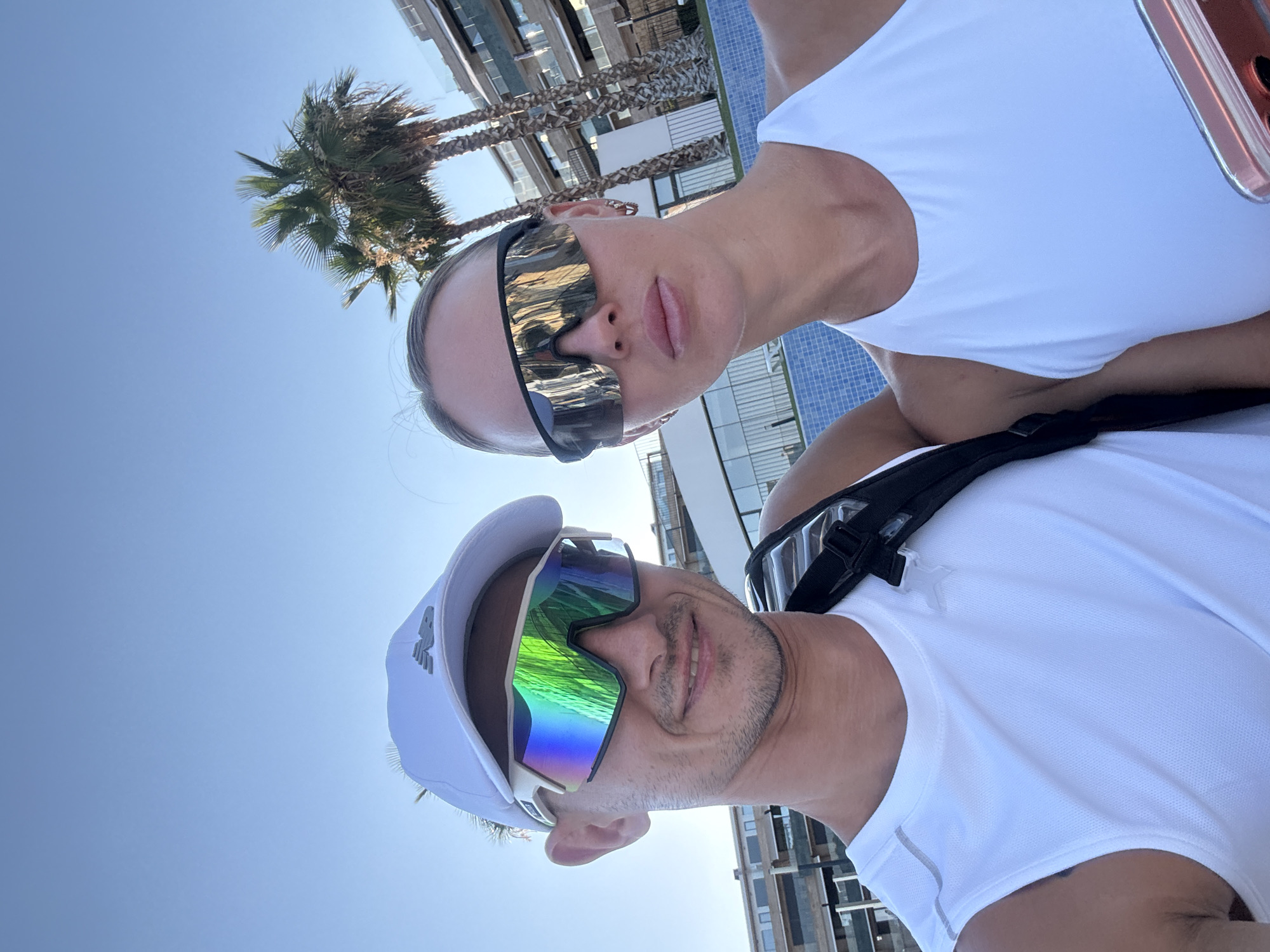 A man and a woman are taking a selfie in front of a building and palm trees. The man is wearing a white baseball cap, white sleeveless shirt, and reflective sunglasses. He has stubble and is smiling. The woman is wearing a white halter top and black-rimmed sunglasses. She has blonde hair pulled back and is looking at the camera. Behind them, a multi-story building with balconies and a swimming pool with blue tiles are visible. Two palm trees stand in the background, and the sky is clear and blue. The scene appears to be in Finestrat, Spain, likely a resort area given the architecture and vegetation.