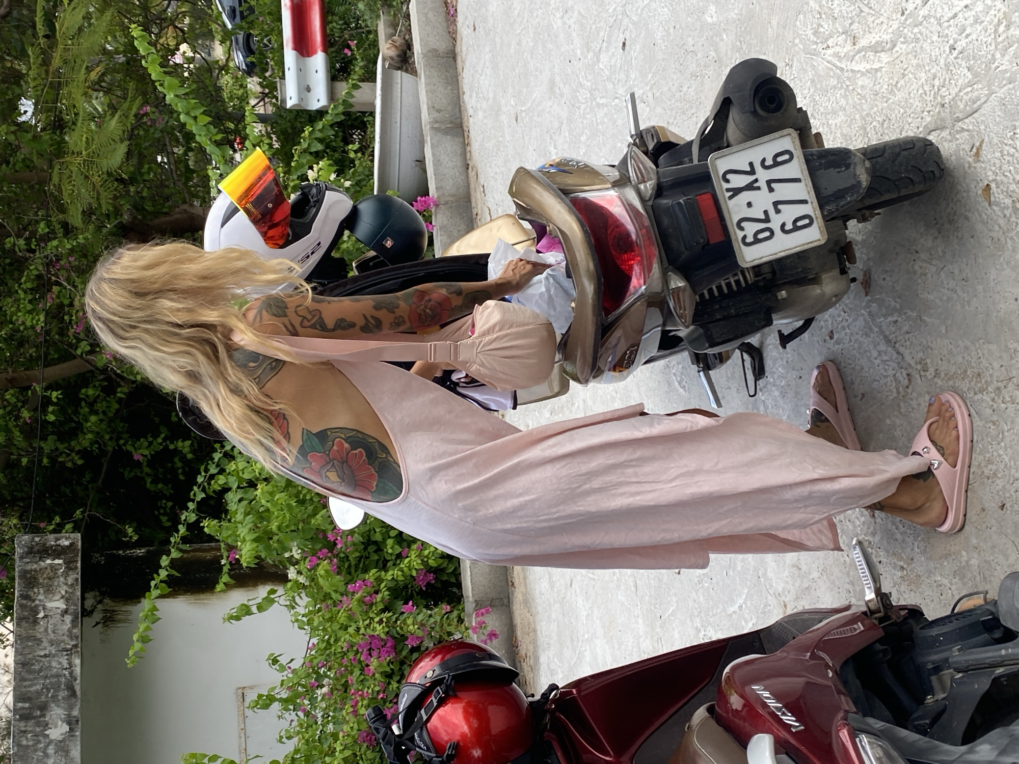 The image captures a blonde-haired woman standing beside a tan/gold scooter in an outdoor setting in Phu Thuy Ward, Vietnam. She wears a light pink sleeveless jumpsuit and matching pink slide sandals adorned with heart embellishments. Her arms and upper back are covered in tattoos, notably a large floral design on her left shoulder blade. The woman appears to be leaning towards the scooter's seat, possibly placing or retrieving items from storage.
The scene features a concrete ground surface and lush green foliage with vibrant pink flowers in the background, characteristic of a tropical or subtropical climate. A red and white striped pole and a concrete pillar are also visible. Two scooters are present: the tan/gold one she is next to, which has a visible license plate reading "62-X2 6776", and a maroon scooter, branded "VISION", partially seen in the foreground. A white full-face helmet with an orange visor (displaying "52"), a dark green/black open-face helmet, and a red open-face helmet are also in view. The image is taken during daytime under bright, sunny conditions.