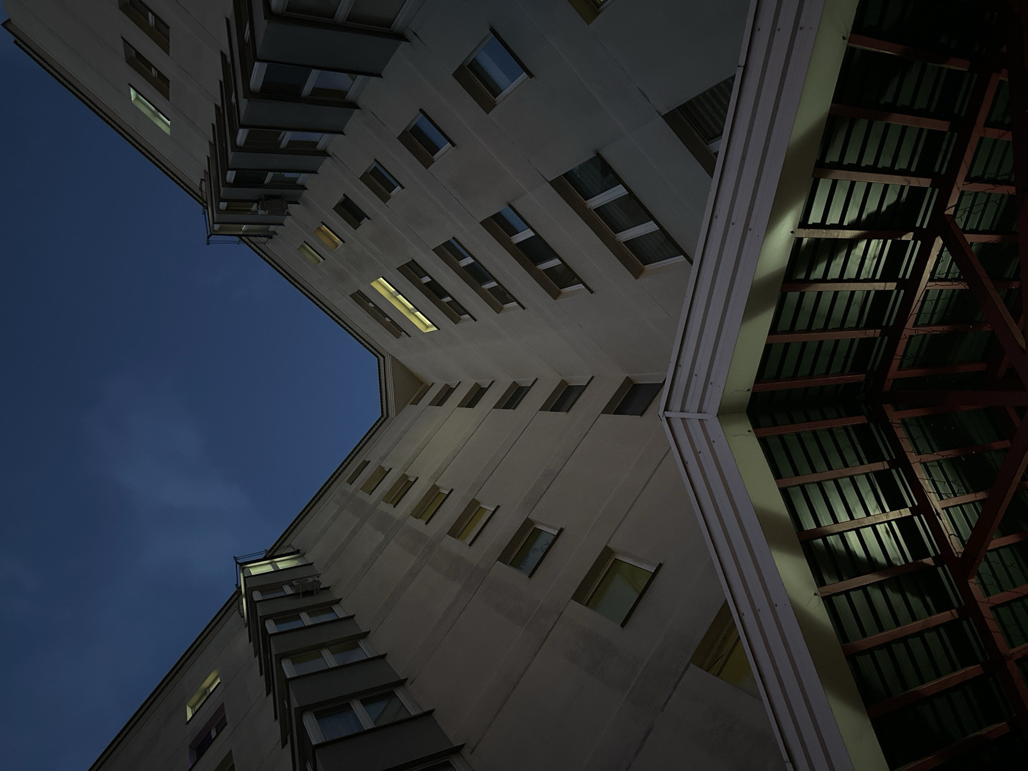 An upward-angle, low-light photograph captures the facade of a multi-story building against a deep blue twilight sky. The building is symmetrical, with two wings angling upwards from a central point, creating a V-shape. Many windows dot the pale beige exterior, some illuminated from within with a yellowish light, others dark. The architectural style appears to be Soviet-era housing. In the foreground, a slanted, covered structure with a green corrugated metal roof and exposed wooden beams is visible, suggesting an entrance canopy or overhang. The overall impression is one of an urban residential environment at dusk in the Ждановіцкі сельскі Савет, Belarus.