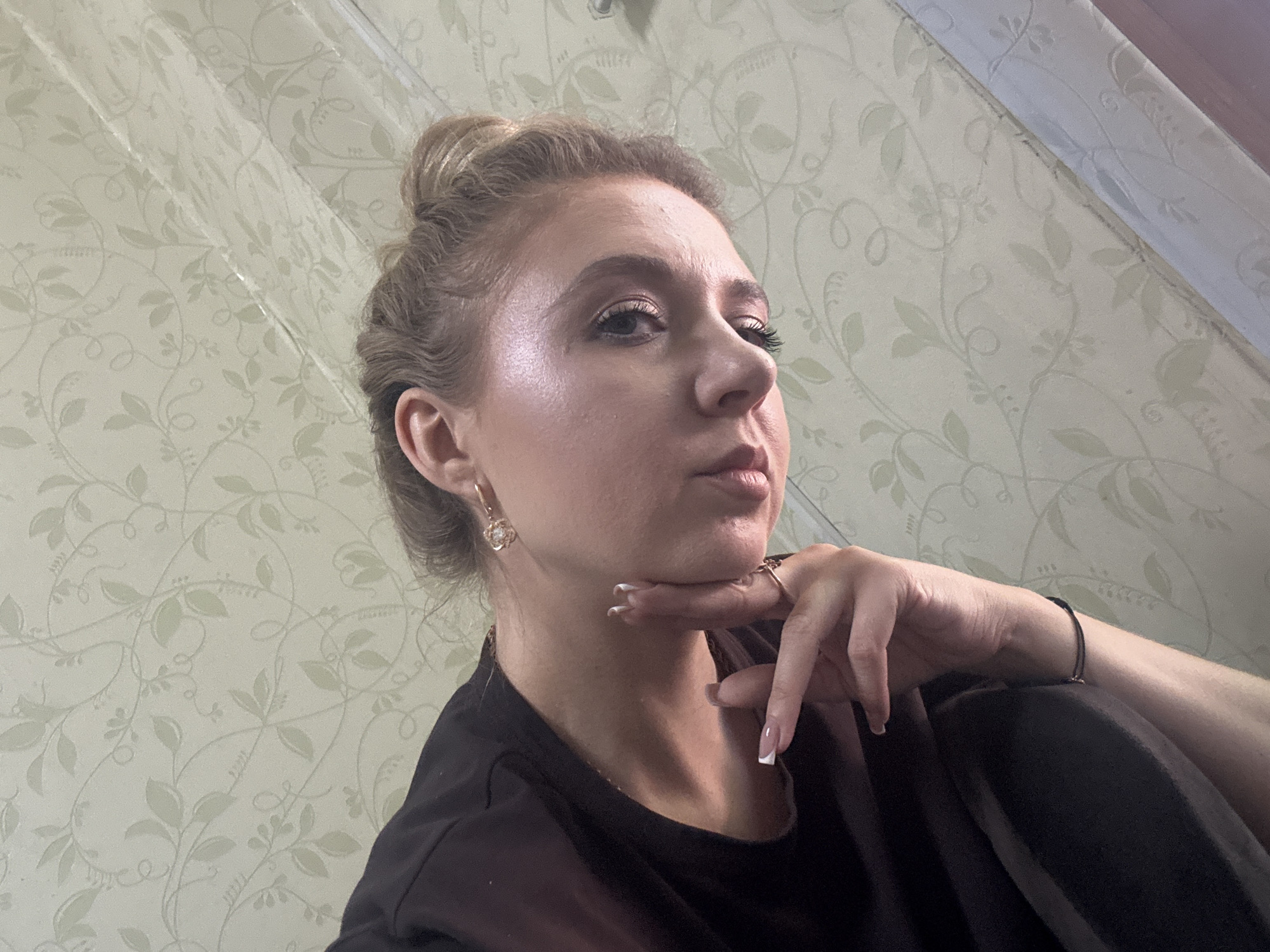 A young woman with blonde hair pulled into a bun is shown in a close-up, medium-shot. She is looking to her right, with her chin resting on her hand. Her left hand is bent, with her fingers touching her chin. Her right arm is bent, with her hand resting on her leg. She is wearing a black shirt. She has on earrings and a ring on her left hand. Her nails are painted white with a French tip. The background is a wall with a floral pattern wallpaper. The lighting suggests it is daytime, and the setting appears to be indoors. The woman's expression is thoughtful or contemplative. No text is visible in the image.