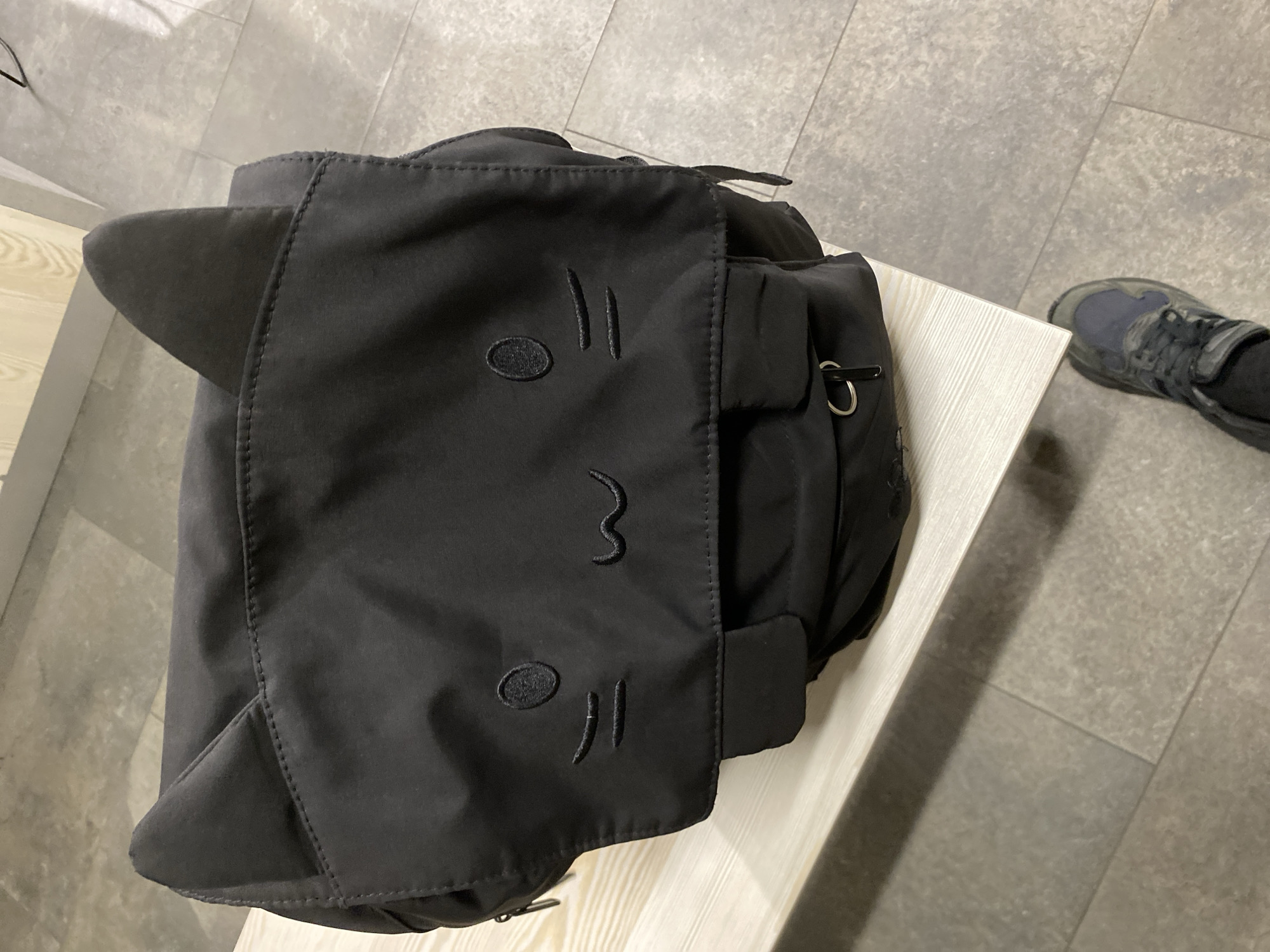 A black cat-themed backpack sits on a light-colored wooden table or bench. The backpack features cat ears on the top corners and an embroidered cat face on the front flap. In the background, there's a tile floor and a foot wearing a gray and black athletic shoe. No specific location information is discernable from the image that would indicate the scene is in Gijón/Xixón, Spain.