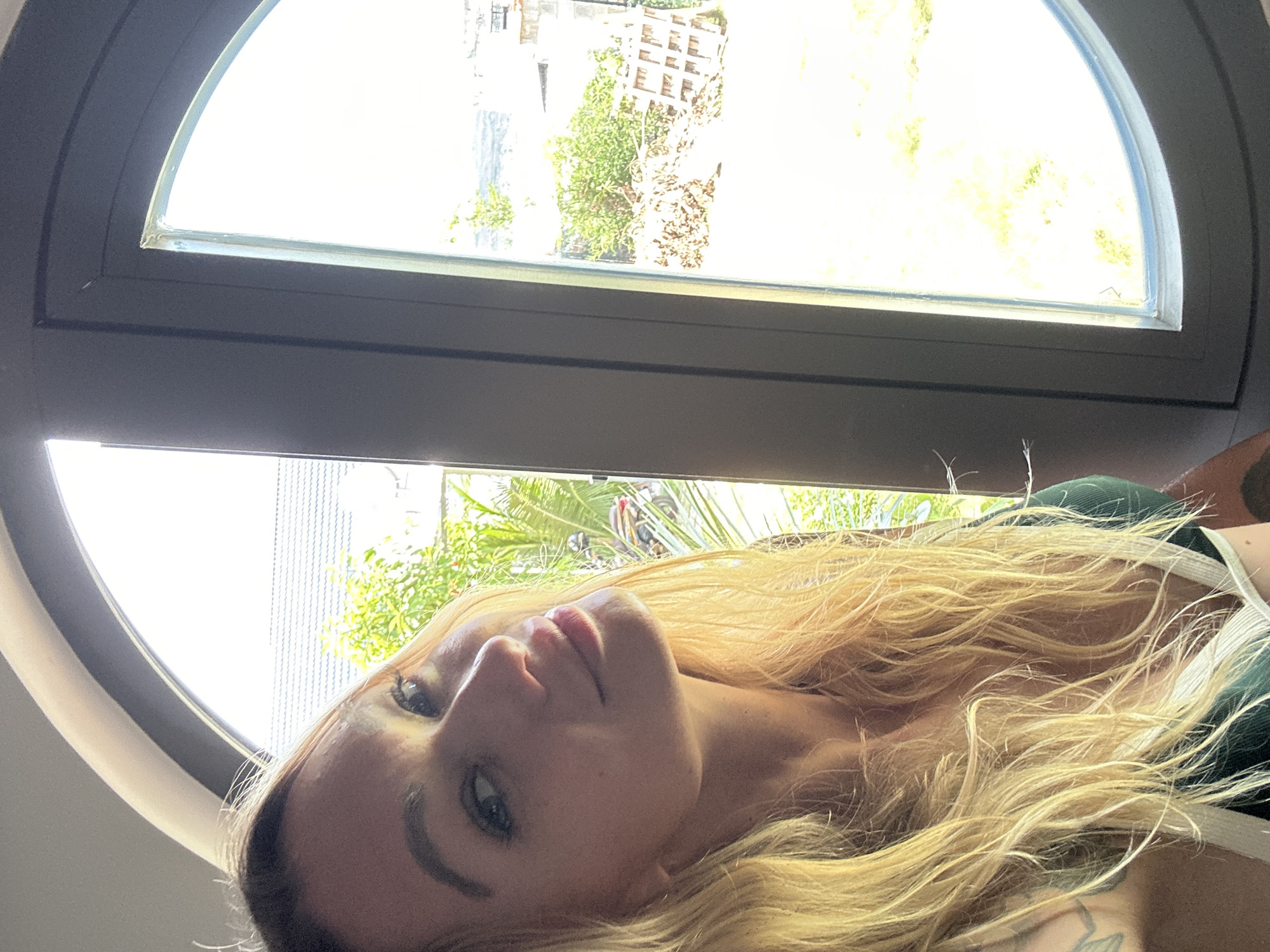 A woman with blonde hair is taking a selfie indoors, next to a large, circular window. She is wearing a dark green top with white trim. The window reveals a bright, sunlit outdoor scene with lush greenery, trees, and some structures in the distance, suggestive of a residential area in Mũi Né, Vietnam. The natural light coming through the window illuminates her face and hair.